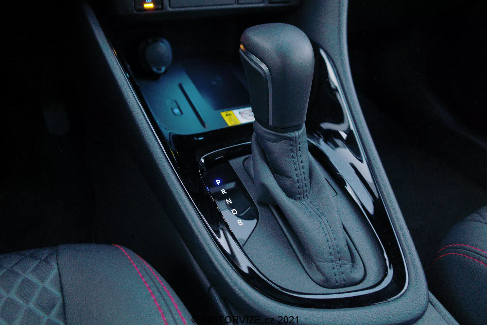 Detailný pohľad na automatickú prevodovku modelu Toyota Yaris 2020, viditeľný zo strany spolujazdca pod miernym sklonom nadol, ktorý zvýrazňuje elegantný kovový povrch a ergonomický dizajn. Prevodovka je umiestnená uprostred medzi sedadlami vodiča a spolujazdca a zahŕňa polohy Park, Reverse, Neutral a Drive, ako aj možnosti manuálneho preraďovania (+ a -). Pred prevodovkou sú elektronické ovládače pre jazdné režimy a kontrolu trakcie spolu s modro osvetleným tlačidlom štartu. Tmavá, lesklá stredová konzola pridáva moderný luxus, doplnený ovládaním vyhrievania sedadiel a výrazným šitím na vodičovom sedadle, čo naznačuje zvýšený komfort a pokročilé jazdné dynamiky.