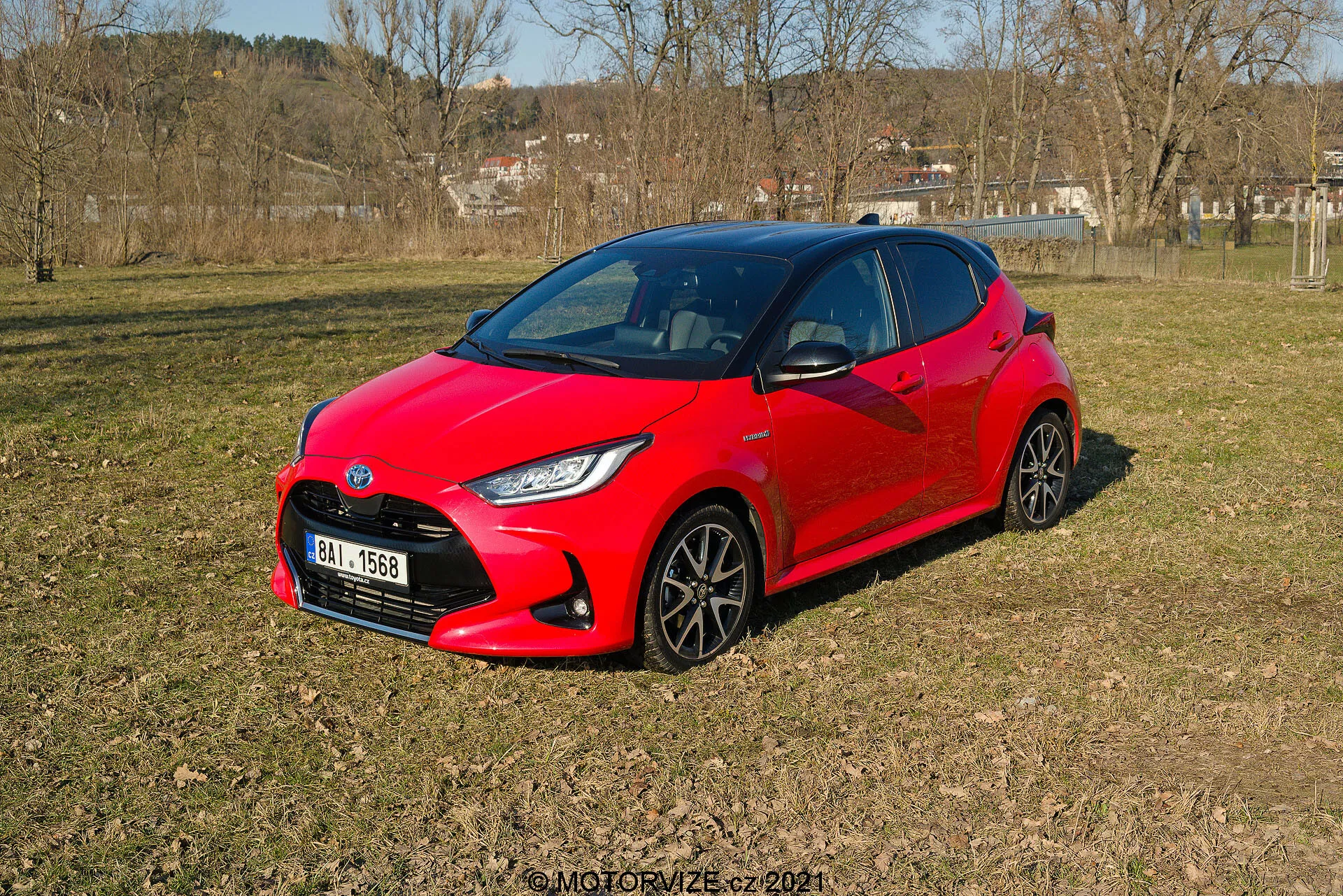 Obrázok zobrazuje žiarivo červený Toyota Yaris 2020, zaparkovaný na nerovnom trávnatom teréne v polovenkovskej oblasti, pohľad z trojštvrťovej perspektívy. Moderný, aerodynamický dizajn vozidla obsahuje ostré uhly okolo svetlometov a kompaktný, športový vzhľad. Viditeľné sú tmavé viaclúčové kolesá a štýlové LED svetlomety. Bočné okná a predné čelné sklo poskytujú tmavý pohľad do interiéru, čo naznačuje možné pokročilé automobilové systémy.