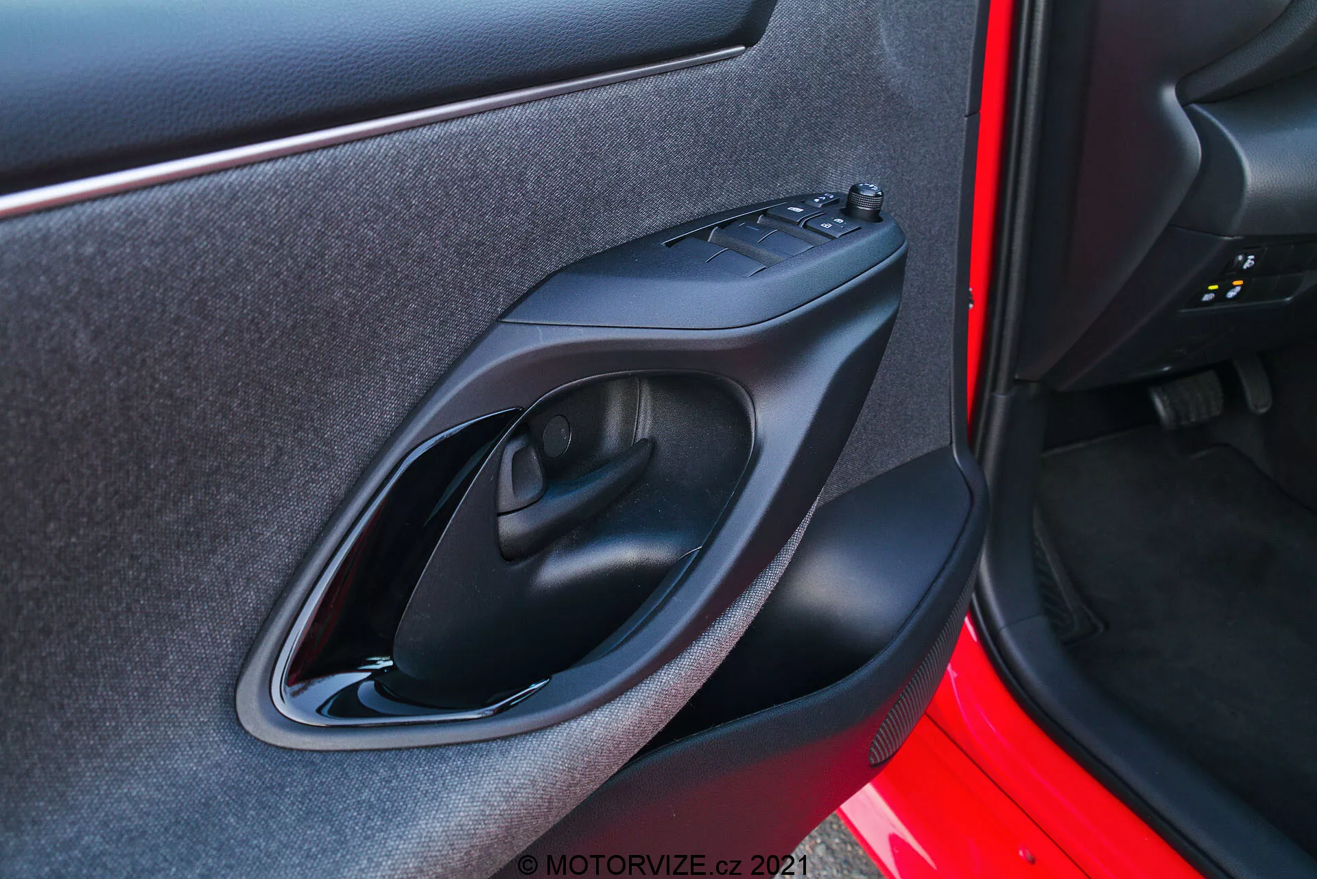 Detailný záber na vnútorný panel dverí vozidla Toyota Yaris 2020 zhotovený z exteriéru. Obrázok sa sústredí na pravú stranu dverového panelu s dizajnovými a technologickými prvkami, ako je matná čierna plastová kľučka dverí, ovládací panel pre elektrické okná, ergonomicky tvarovaná opierka na ruku a malé úložné vrecko. Vonkajšia strana dverí je natretá živou červenou farbou.