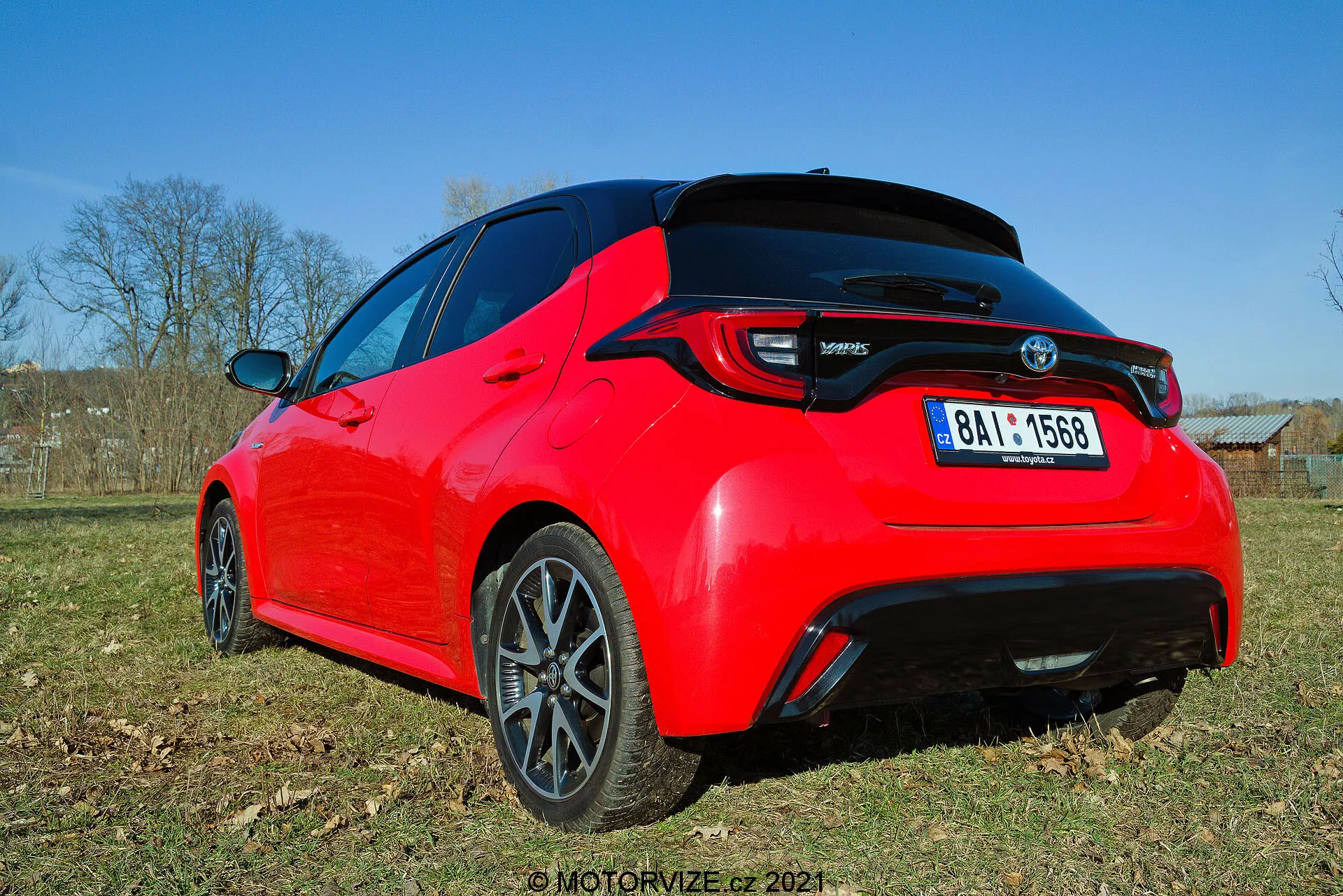 Zadný trojštvrťový pohľad na jasne červený hatchback Toyota Yaris 2020, videný z nízkeho uhla, zvýrazňujúc dizajnové línie a kľúčové prvky ako sú LED zadné svetlá, zadný spojler, anténa v tvare žraločej plutvy a tmavé viacramenné zliatinové disky. Fotografia zachytáva detail zadnej časti a čiastočne bočný profil na dennom svetle, zvýrazňujúc športovú estetiku a moderné prvky.