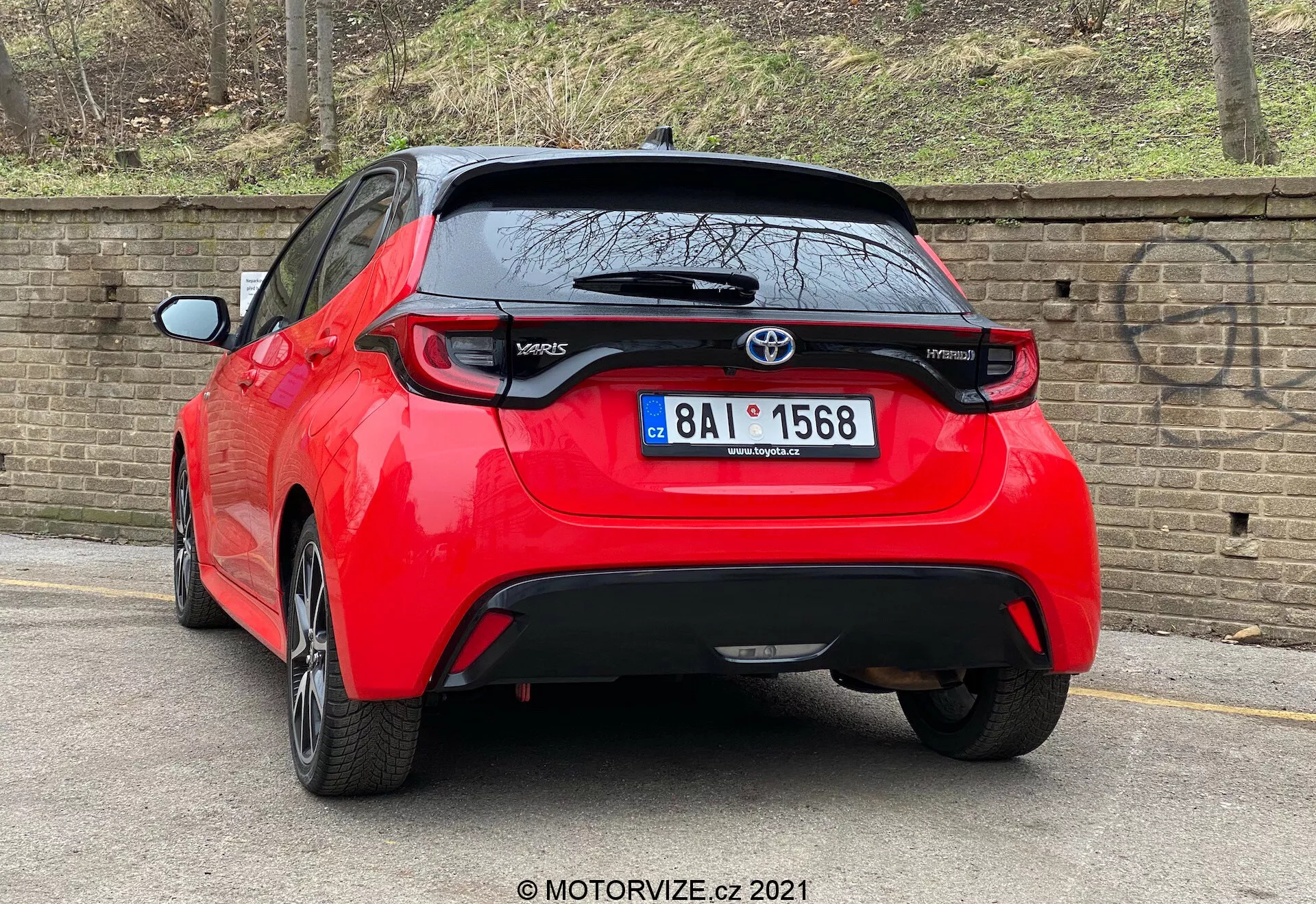 Vysokokvalitný obrázok červeného hatchbacku Toyota Yaris 2020 zobrazeného zo zadného trojštvrťového uhla. Vozidlo má charakteristické zadné svetlá, centrálny emblém Toyota nad pravdepodobnou zadnou kamerou a označenie 'Yaris' na spodnej ľavej strane zadných dverí. Pod oblasťou registračnej značky sa nachádza čierny ozdobný prvok, ktorý by mohol byť vyrobený z plastu alebo uhlíkových vlákien, pričom malý zadný spojler presahuje zadné okno. Auto, zaparkované blízko obrubníka na dláždenej ceste vedľa tehlového múru, naznačuje mestské alebo rezidenčné prostredie.