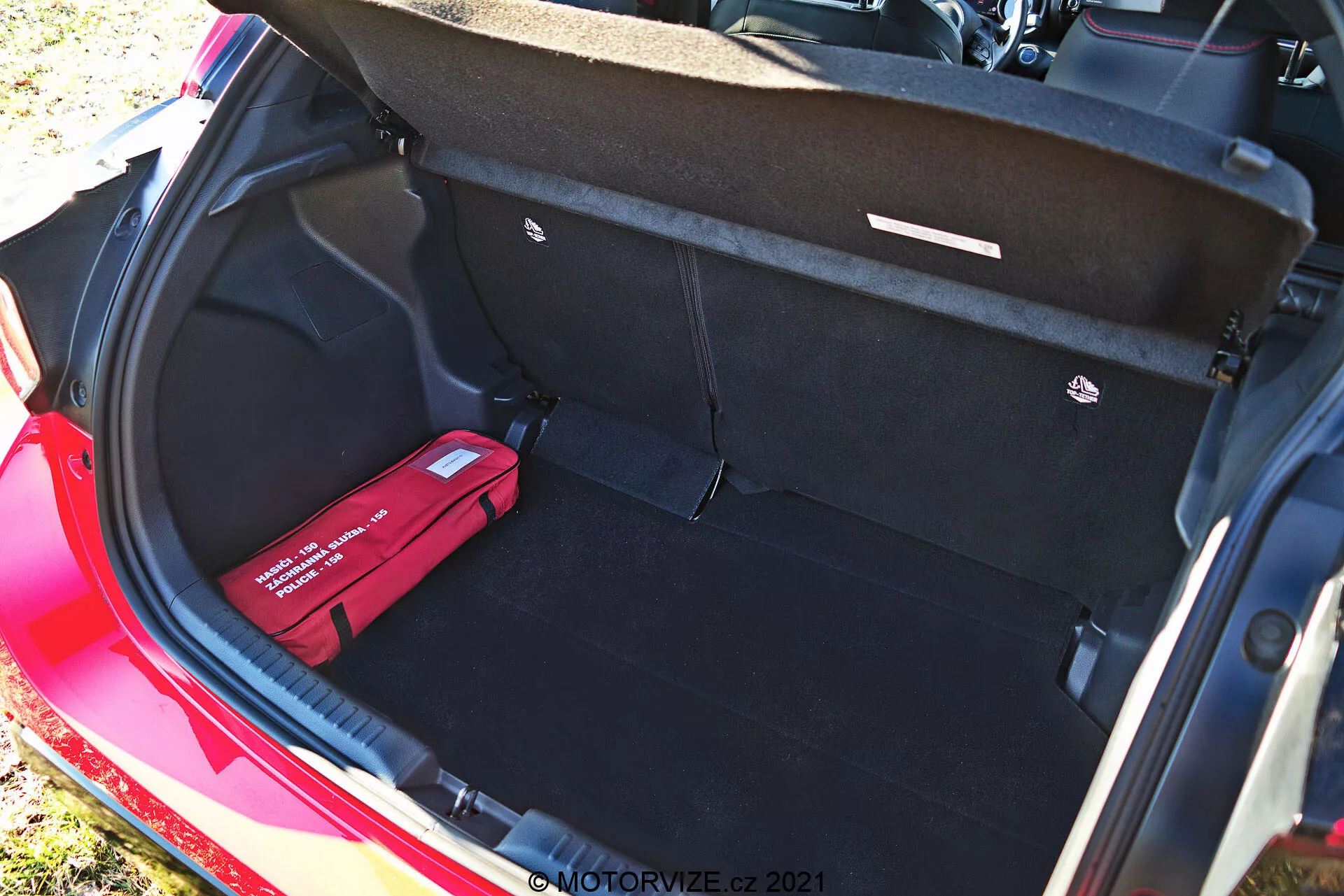Batožinový priestor červeného Toyota Yaris 2020 z pohľadu z vyššieho uhla, ktorý ukazuje jeho čistý, priestranný interiér s čiernym kobercom, otvoreným krytom batožinového priestoru s ťahadlami, plochou nakladacou podlahou, hákom na batožinu napravo a červenou lekárničkou alebo núdzovou súpravou vľavo. Táto denná fotografia zvýrazňuje dostatočnú úložnú kapacitu a organizačné prvky vozidla.