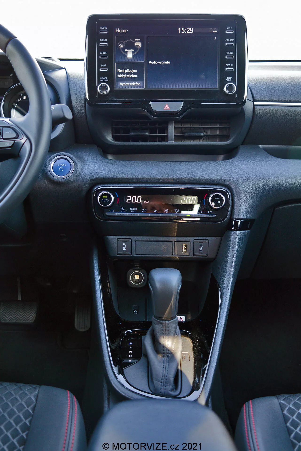 Pohľad na interiér Toyota Yaris 2020 sústredený na stredovú konzolu a prvky prístrojovej dosky. Fotografia, urobená z predného sedadla spolujazdca, zobrazuje digitálny infotainment systém s dotykovou obrazovkou, zobrazujúci domovskú obrazovku s časom 15:29 a rôznymi možnosťami menu, centrálne ventilácie a používateľsky prívetivé ovládače klimatizácie a vykurovania s digitálnym displejom teploty nastavenou na 20,0 stupňov. Stredová konzola obsahuje koženú radiacu páku so strieborným akcentom a ručnú parkovaciu brzdu. Interiér je navrhnutý v tmavých tónoch s kovovými úpravami, poskytujúc moderný a sofistikovaný vzhľad.