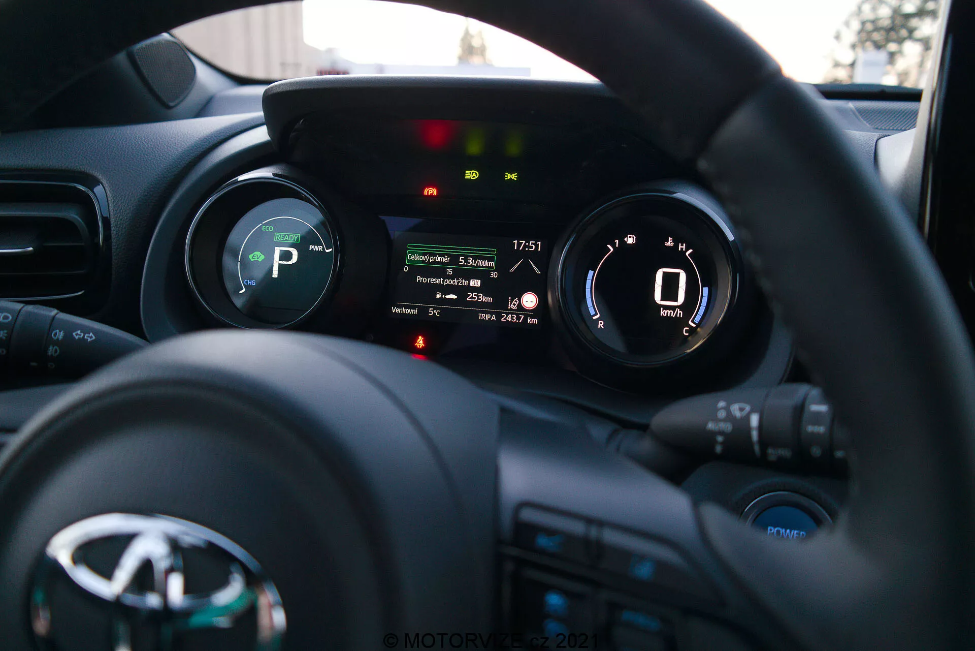 Fotografia interiéru vozidla Toyota Yaris 2020 z pohľadu vodiča, so zameraním na volant a prístrojovú dosku. Volant s logom výrobcu a viacerými ovládacími tlačidlami pre médiá, komunikáciu a tempomat je v dolnej časti. Prístrojový panel za volantom obsahuje digitálny displej so zobrazením rýchlomeru ukazujúceho 0 km/h a indikátory pre prevodový stupeň (P ako Parkovanie), pripomienku bezpečnostného pásu, upozornenie na otvorené dvere a ďalšie svetlá stavu vozidla. Malá hĺbka ostrosti zvýrazňuje prístrojový panel, zatiaľ čo prístrojová doska a interiér vozidla sú jemne rozmazané na pozadí. Obrázok pravdepodobne zhotovený neskoro popoludní alebo skoro večer ukazuje modernú prístrojovú techniku ​​a technológiu rozhrania vozidla.