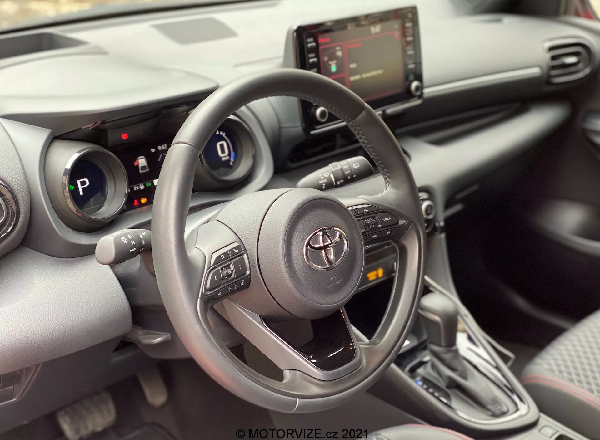 Pohľad na interiér Toyoty Yaris 2020 z miesta spolujazdca, zameraný na oblasť vodiča. Obrázok zvýrazňuje trojramenný volant Toyota s ovládacími tlačidlami pre audio, Bluetooth a tempomat, s emblémom Toyota uprostred. Za volantom je čiastočne viditeľný digitálny prístrojový panel, ktorý ukazuje, že auto je v režime „P“ (parkovacie). Stredová konzola zobrazuje infotainment obrazovku a pod ňou ovládanie HVAC, spolu s možnými USB alebo aux vstupmi. Obrázok tiež ukazuje automatickú prevodovku s moderným dizajnom a krytom z materiálu podobného koži. Interiér má moderný dizajn s kombináciou mäkkých materiálov a strieborných akcentov, spolu s držiakmi na nápoje alebo malými úložnými priehradkami vedľa radiacej páky. Čiastočne viditeľný panel dverí odhaľuje ovládací panel okien, čo naznačuje elektrické okná.