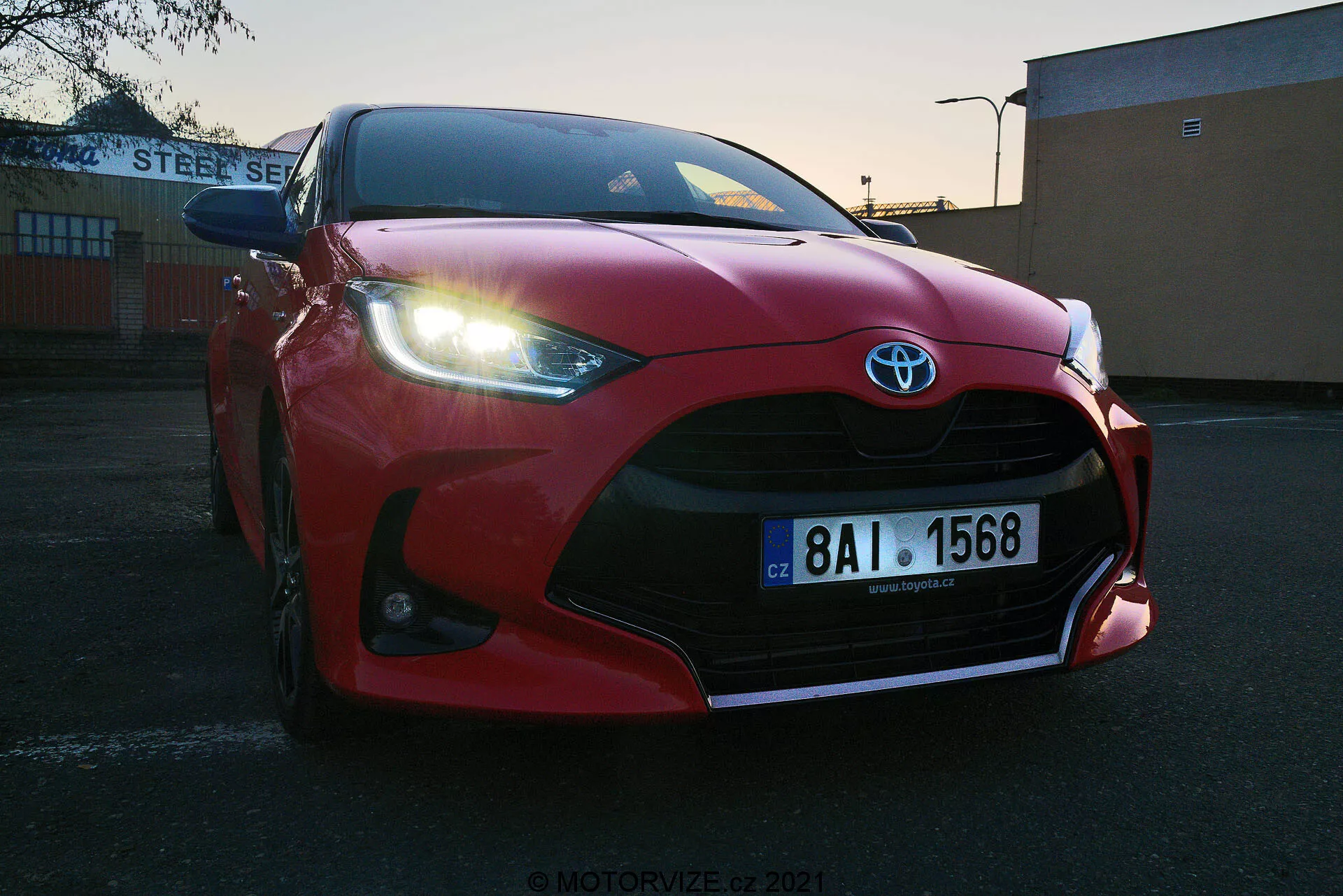 Nízky uhol zblízka červeného vozidla Toyota Yaris 2020 zaparkovaného na vonkajšom parkovisku pri východe alebo západe slnka. Zdôraznená predná mriežka, nárazník, aerodynamicky tvarovaná kapota, ostré LED svetlomety a veľké vzduchové otvory zvýrazňujúce znak Toyota. Viditeľná európska poznávacia značka, umiestnená na pozadí priemyselnej alebo komerčnej budovy s nápisom 'STEEL SERVICE'. Jasná obloha s prechodovým svetlom zvýrazňuje športový dizajn vozidla.