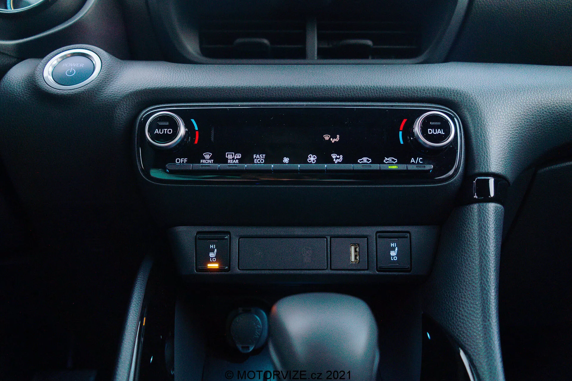Detailný pohľad na palubnú dosku Toyota Yaris 2020: Obrázok zdôrazňuje klimatizačný systém s dvojzónovou automatickou klimatizáciou, s tlačidlami 'AUTO' a 'DUAL' pre nezávislé nastavenie teploty pre vodiča a spolujazdca. Pod ovládacími prvkami klimatizácie je viditeľný úložný priestor s USB portom a potenciálnou 12V zásuvkou. Nižšie sú viditeľné tri ovládače: centrálny je radiaca páka v polohe 'Park', ktorá naznačuje automatickú prevodovku, menší otočný ovládač naľavo pravdepodobne na výber režimu jazdy a tlačidlo napravo, ktoré môže byť elektronická parkovacia brzda. Materiál palubnej dosky má tmavú farebnú schému s prémiovým textúrovaným povrchom, čím vytvára moderné, používateľsky prívetivé prostredie kabíny.