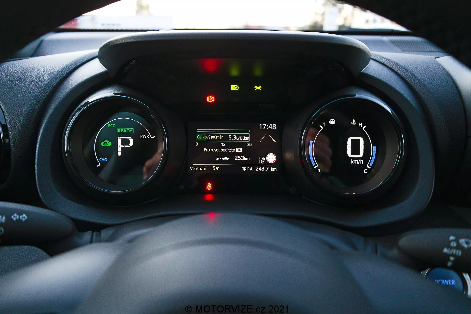 Pohľad do interiéru Toyota Yaris 2020 zameraný na prístrojový panel, urobený z pohľadu vodiča, zobrazujúci digitálne prístroje, indikátory elektrického alebo hybridného vozidla a multifunkčný displej s funkciami ako vzdialenosť cesty, vonkajšia teplota a dojazd batérie.