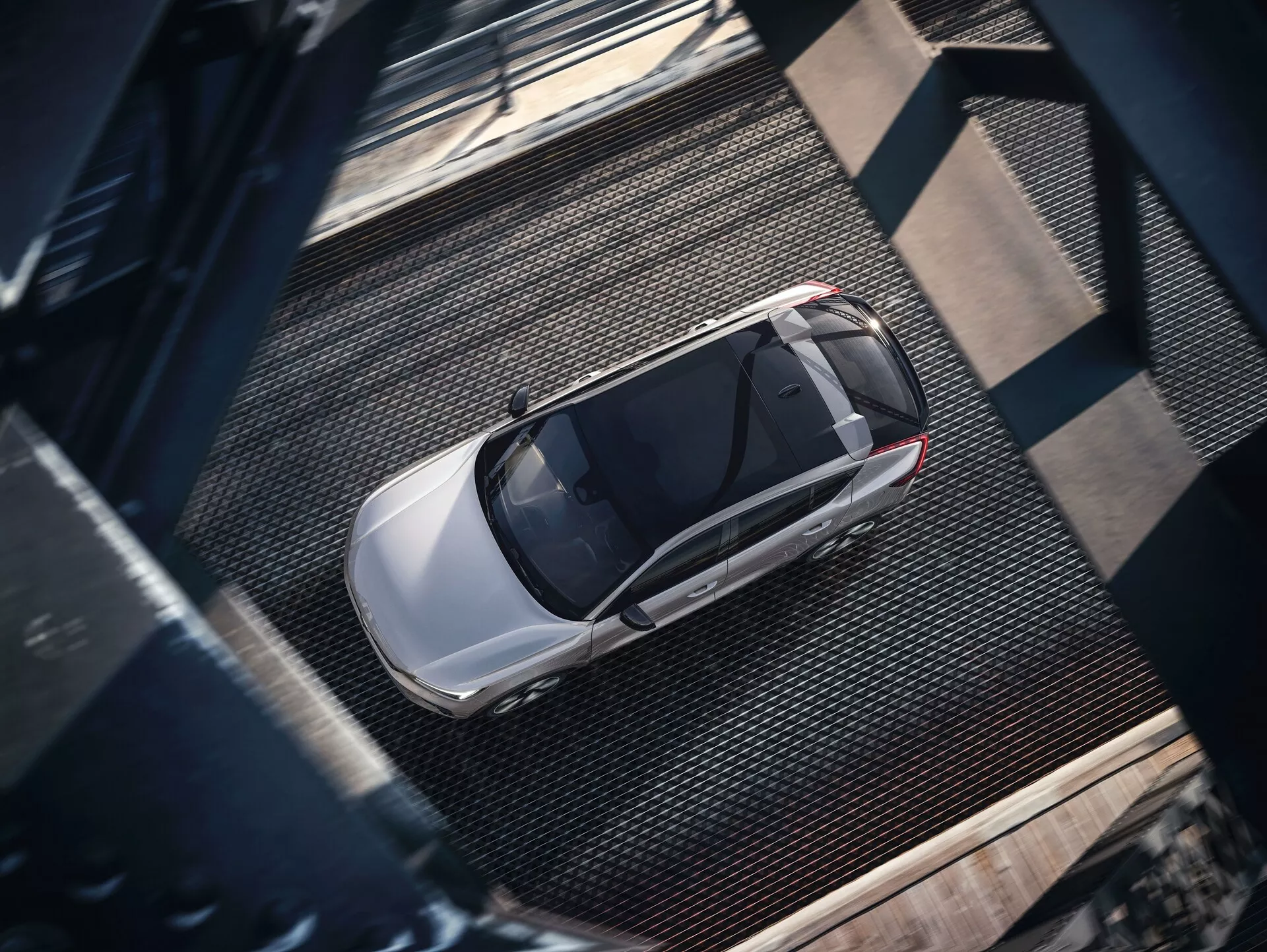 Pohľad z vtáčej perspektívy na Volvo C40 2021 prvej generácie, zachytený z výšky. Obrázok odhaľuje úplný horný pohľad na elegantný, moderný sedan s ľahko metalickým lesklým povrchom, pravdepodobne strieborne alebo svetlosivý. Viditeľné prvky zahŕňajú strechu, kapotu, kufor, okná a časti dverí s výrazným strešným oknom naznačujúcim pokročilé vetranie. Auto je zaparkované na textúrovanom priemyselnom povrchu s mriežkovými textúrami a tmavými nitovanými nosníkmi, vrhajúcimi tiene naznačujúce bočné osvetlenie pravdepodobne ráno alebo neskoro popoludní.