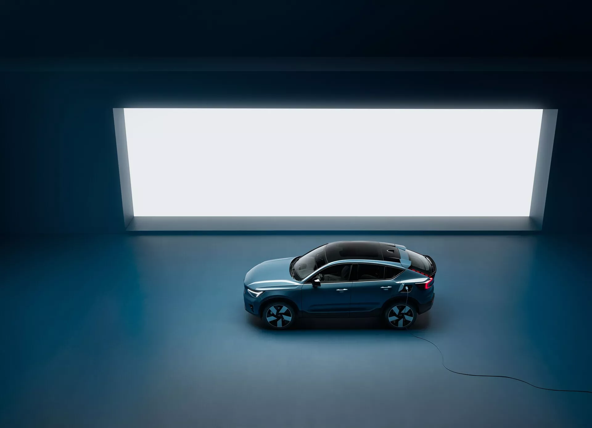 Záber z vysokého uhla na sedan Volvo C40 2021 so štyrmi dverami a panoramatickým strešným oknom, pohľad zhora na tmavú podlahu s jasným obdĺžnikovým svetelným zdrojom v pozadí, ktorý ukazuje čistý a minimalistický dizajn s lesklou tmavou karosériou a veľkými viacramennými ľahkými kolesami, zdôrazňujúcimi luxusný a moderný vzhľad.