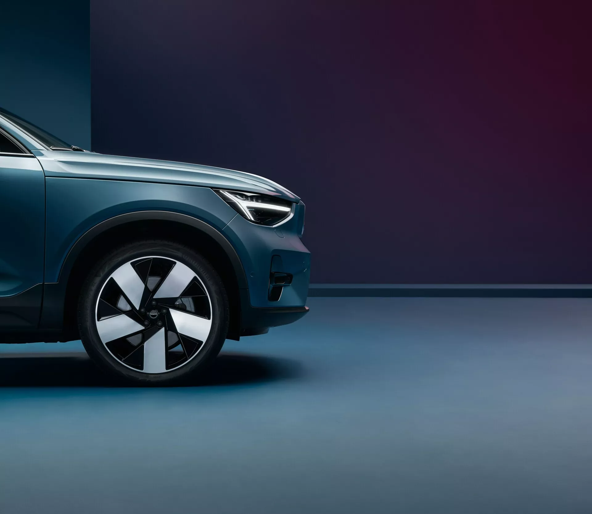 Nízky uholný záber na Volvo C40 2021 (prvá generácia), zameriavajúci sa na prednú časť od predného kolesa po stred kapoty. Zvýraznené sú moderné svetlomety, viacramenný zliatinový disk, metalický lesklý lak a aerodynamické línie. Dvojfarebné pozadie s jemným osvetlením podtrhuje prémiovú úpravu a silnú postavu vozidla.