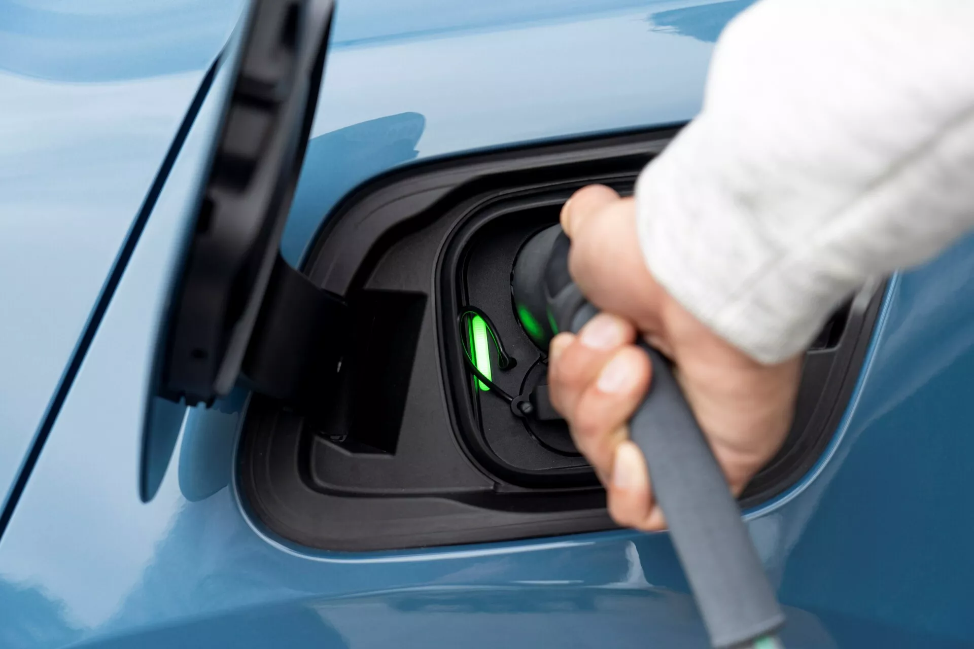 Zblízka bočný pohľad na oblasť nabíjacieho portu elektrického vozidla Volvo C40 z roku 2021 (1. generácia), s ľudskou rukou pripojujúcou nabíjací kábel k portu. Zameranie na proces nabíjania, zdôrazňuje otvorený nabíjací port s pravdepodobne použitým konektorom typu 2. Zelené svetlo vo vnútri zásuvky signalizuje, že vozidlo je pripravené na nabíjanie alebo sa aktuálne nabíja. Pohľad je mierne zvýšený, ukazuje bezchybný modrý lak vozidla, zdôrazňujúc čistý a moderný aspekt elektrického vozidla.