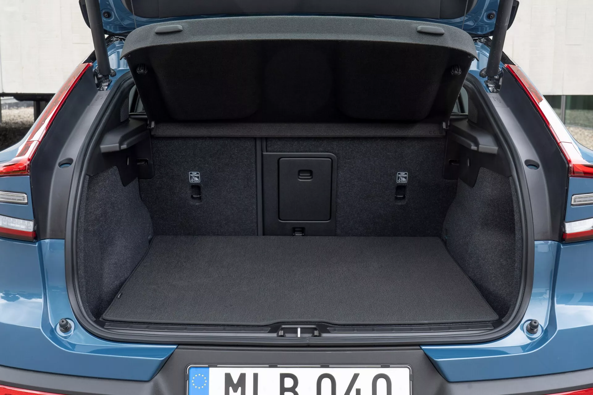 Zadný batožinový priestor moderného auta, ukazujúci kufor Volvo C40 2021 (1. generácia) z pohľadu zozadu, mierne pod úrovňou pása. Veko kufra je otvorené, zvýrazňujúc štandardné polstrovanie vnútorného obloženia. Kufor je prázdny, zobrazuje vnútorný koberec, zapustené bočné oblasti a rukoväť v strede podlahy, ktorá umožňuje prístup do úložného priestoru. Nie sú viditeľné žiadne háky na batožinu ani upevňovacie body. Európsky štýl poznávacej značky naznačuje európsky pôvod.