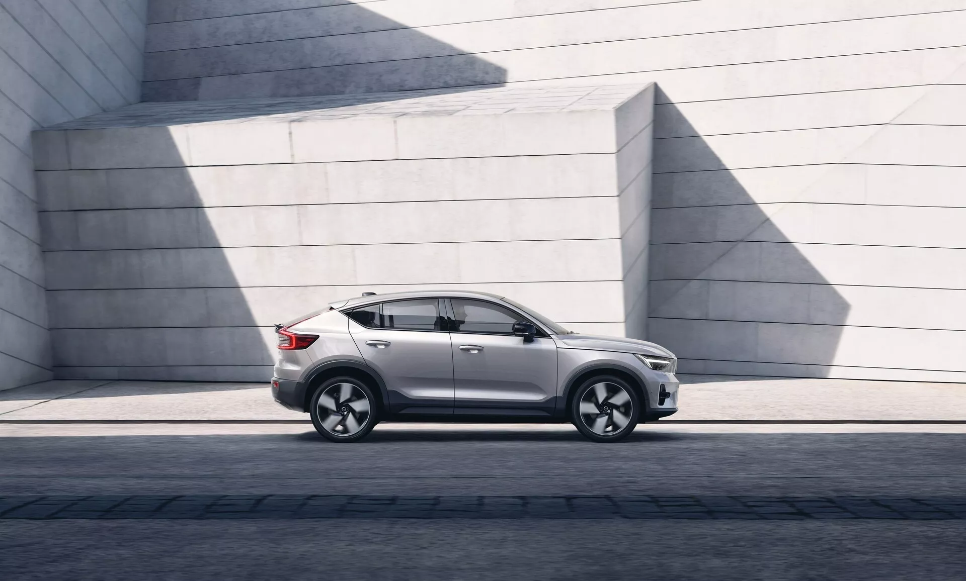 Obrázok moderného SUV Volvo C40 2021 zaparkovaného paralelne, ukazujúci ľavý bočný profil od ľavého predného svetlometu po ľavé zadné svetlo. Uhol zdôrazňuje svetlú výšku a postoj na dláždenom povrchu. Slnko vytvára tiene na lesklom lakovanom povrchu. SUV má elegantný dizajn s mierne šikmou strechou, výraznými kontúrami dverí, tónovanými oknami a modernými bočnými zrkadlami. Kolesá z ľahkých zliatin s viacramenným dizajnom a nízkoprofilovými pneumatikami naznačujú výkon a luxus. Viditeľné sú bočné prahy, tvarovanie blatníkov a charakterová línia prebiehajúca cez kľučky dverí, naznačujúca robustnosť. Neprítomnosť viditeľného výfuku naznačuje možný elektrický alebo hybridný pohon. Minimalistické betónové štruktúry v pozadí kontrastujú s jemnými krivkami auta, zdôrazňujúc jeho mestský a dizajnérsky vzhľad.