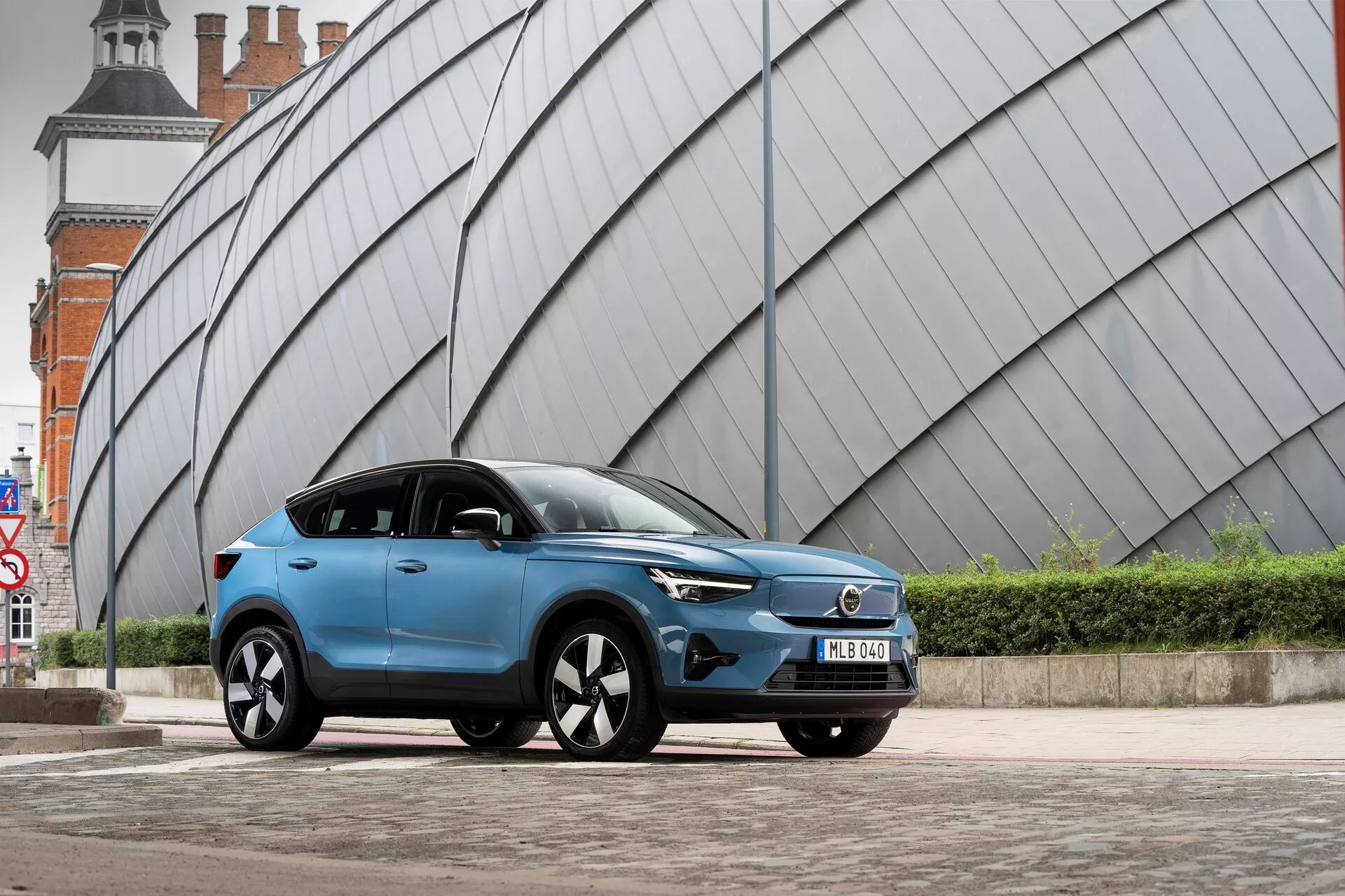 Moderné modré SUV Volvo C40 2021 zaparkované na dláždenom mestskom mieste s uhlovou kovovo vyzerajúcou štruktúrou v pozadí. Nízkouhlový záber z ľavej prednej strany ukazuje prednú mriežku vozidla, LED svetlomety, aerodynamický profil, viacramenné zliatinové kolesá, bočné zrkadlá s integrovanými smerovými svetlami, matný alebo saténový povrch farby, zatmavené sklá, prednú poznávaciu značku a sošné bočné línie karosérie.