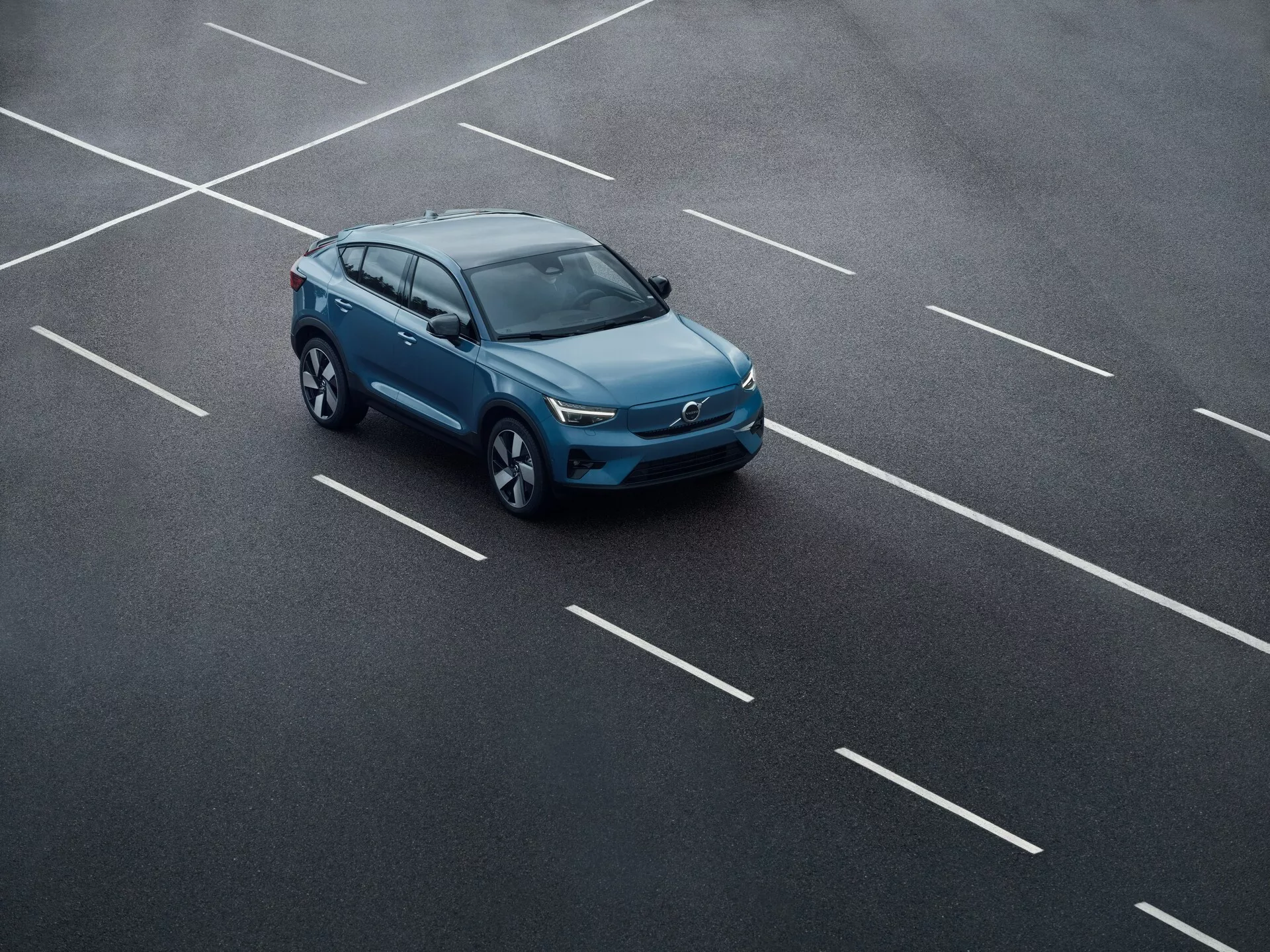 Pohľad zhora na Volvo C40 2021 (1. generácia) na parkovisku, umiestnené diagonálne cez viaceré miesta. Obraz zachytáva horné a bočné profily vozidla, zdôrazňuje strechu, kapotu, bočné zrkadlá, kolesá a hornú mriežku. Elegantný a moderný dizajn, prípadne strešné okno, strešné nosiče a LED svetlomety naznačujú technologické pokroky.