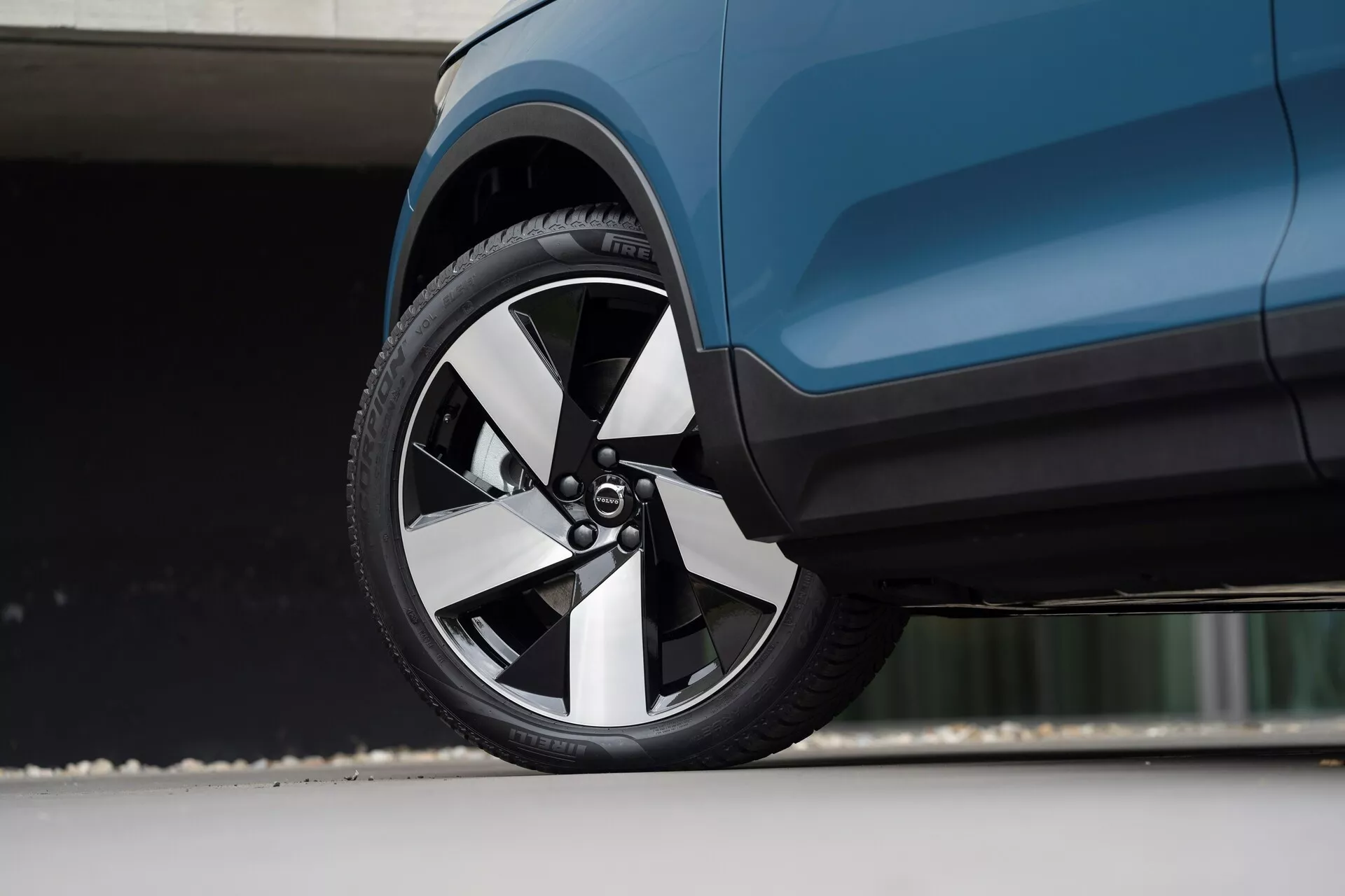 Detailný záber z nízkeho uhla na moderné Volvo C40 2021 (1. generácia), so zameraním na predné koleso a blízku bočnú karosériu. Viacramenný ráfik má kombináciu kovových a tmavších povrchov, doplnenú nízkoprofilovou pneumatikou. Aerodynamická karoséria tmavomodrého vozidla zvýrazňuje čisté línie a sofistikované dizajnové prvky.