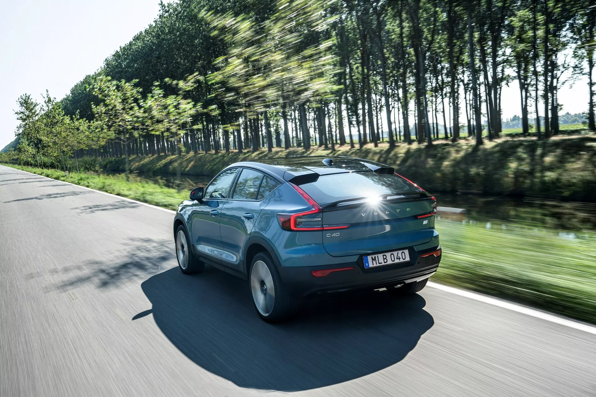 Dynamický záber na Volvo C40 z roku 2021 (1. generácia) v pohybe za slnečného dňa, zadný pohľad z troch štvrtín, ktorý zdôrazňuje elegantný profil a aerodynamiku vozidla. Auto, lakované lesklou modrou farbou, má výrazné zadné LED svetlá, pohybom rozmazané kolesá a stromy odrážajúce jeho rýchlosť. Výrazné detaily zahŕňajú panoramatickú strechu, zadný spoiler a ŠPZ na zadných dverách, čo zdôrazňuje moderné dizajnové prvky a pokročilú osvetľovaciu technológiu.