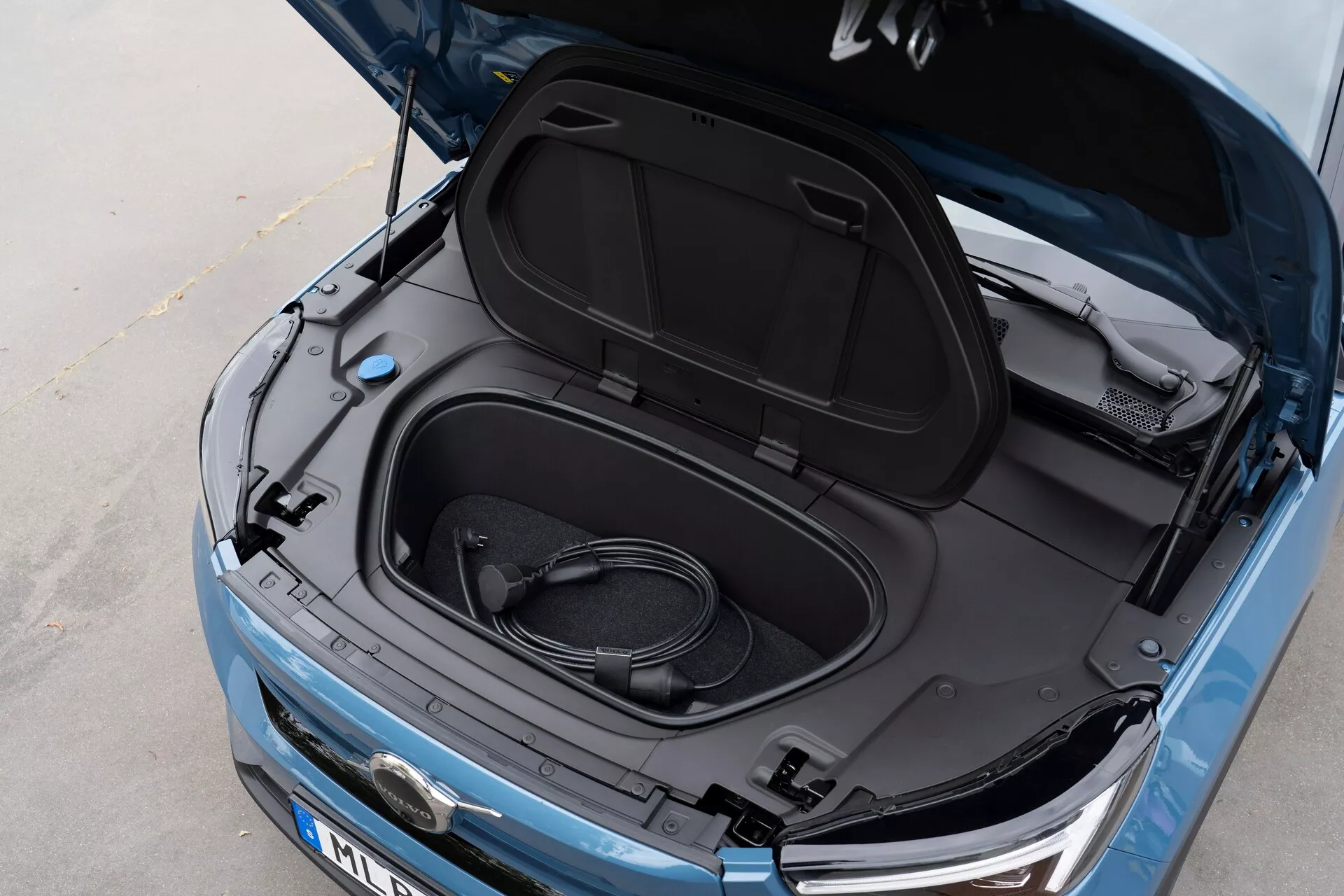 Detailný pohľad zhora z uhla na automobil Volvo C40 2021 (1. generácia) s otvoreným frunkom, ktorý odhaľuje prázdny úložný priestor a zvinutý nabíjací kábel, čo naznačuje, že ide o elektrické vozidlo. Kovovo modrá farba, čierny vnútro veka frunku s informačnými nálepkami, čiastočný pohľad na logo výrobcu, elegantné svetlomety a viditeľná predná mriežka, naznačujúca EV s modernou LED technológiou a aerodynamickým dizajnom.