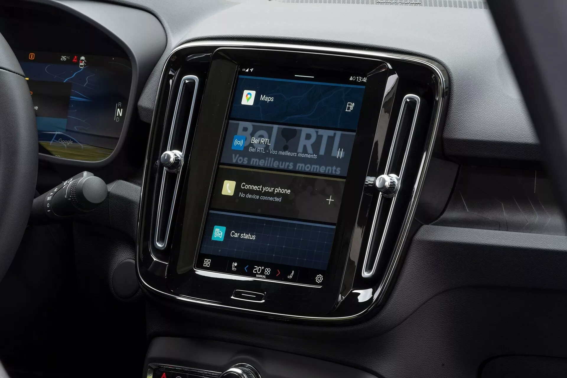 Detailný pohľad na infotainment systém vo vnútri Volvo C40 2021. Záber je urobený z mierne nadol skloneného uhla a sústredí sa na dotykový displej osadený v lesklej čiernej obrube. Displej zobrazuje možnosti ako mapy, médiá, pripojenie telefónu a stav auta, pričom 'Pripojte svoj telefón' je interaktívny prvok. Okolo infotainment systému sú mäkké materiály a obrysové línie naznačujúce vetracie otvory, čo naznačuje moderný interiér auta. Nastavenie teploty ukazuje 20° Celsia a na ľavej strane je viditeľná časť volantu.