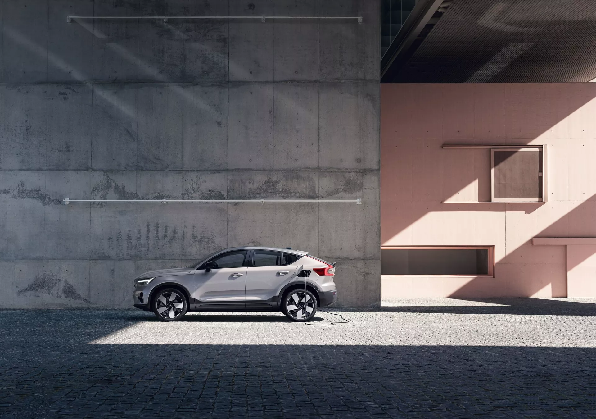 Bočný profil elegantného SUV Volvo C40 z roku 2021 (1. generácia) proti mestskému pozadiu s betónovou a broskyňovou stenou. Auto má reflexný biely lak, štýlové zliatinové ráfiky, tónované okná, strieborné strešné nosiče a integrované smerovky v spätných zrkadlách. Fotografia zvýrazňuje dizajnové línie a krivky vozidla pod prirodzeným denným svetlom, čo pridáva na luxusu a sofistikovanosti.