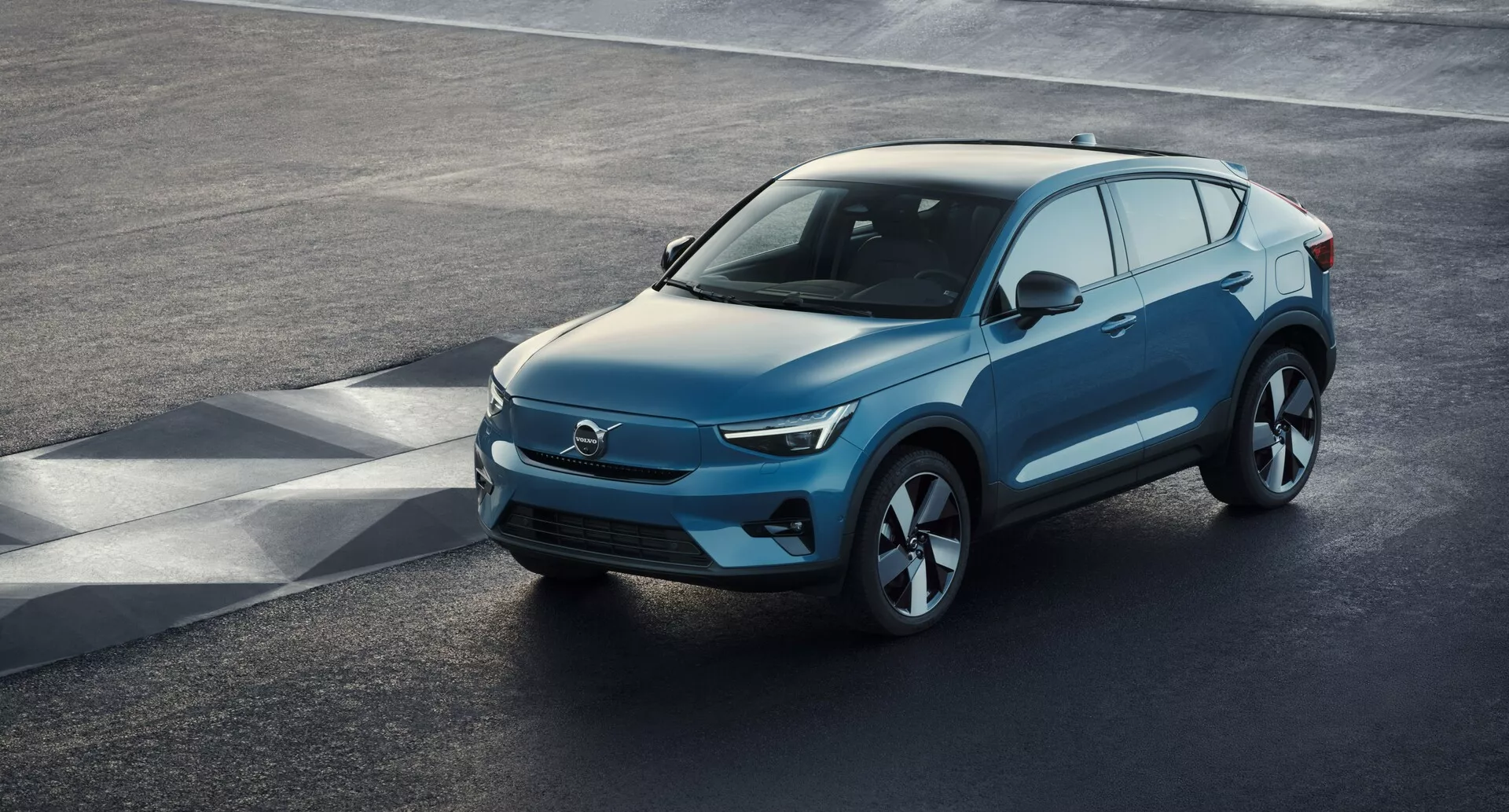 Výhľad z vyvýšeného uhla na moderné SUV Volvo C40 2021 (1. generácia) zaparkované na asfaltovom povrchu, zobrazujúce vysokouhlový trojštvrťový predný pohľad. Elegantná predná časť, pravdepodobne charakteristická pre elektrické vozidlá s minimálnymi prívodmi vzduchu, sa spája s ostrými LED svetlometmi, hladkou zvažujúcou sa kapotou a tvarovaným bočným profilom so veľkými zliatinovými kolesami. Zrkadlá so zabudovanými smerovými svetlami, dostatok okien s možným súkromím a jemne zvažujúca sa línia strechy s nosičmi strechy pre úžitkové vlastnosti sú výrazné rysy. Jasný pohľad zachytáva luxusný dizajn vozidla, pokročilú aerodynamiku a moderné technologické prvky.