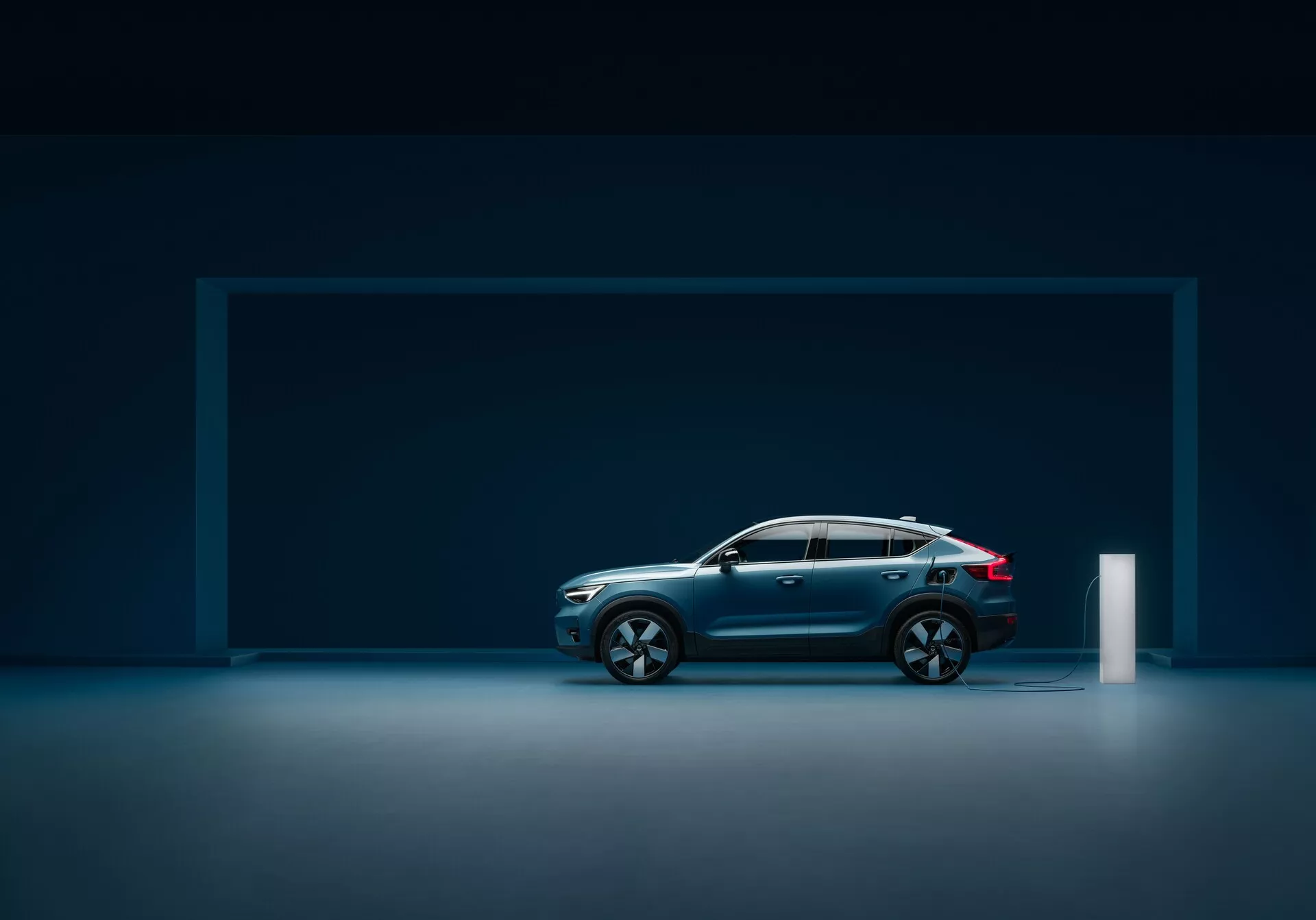 Bočný profil moderného vozidla Volvo C40 2021 (1. generácia) v štúdiovom prostredí s reflexnou podlahou, ukazujúci elegantný dizajn, tmavomodrý metalický lak, klesajúcu líniu strechy, veľké viacramenné zliatinové kolesá, kontúrované línie, robustnú konštrukciu, integrované bočné zrkadlá s indikátormi smeru, LED zadné svetlá a elektrickú nabíjaciu zásuvku.