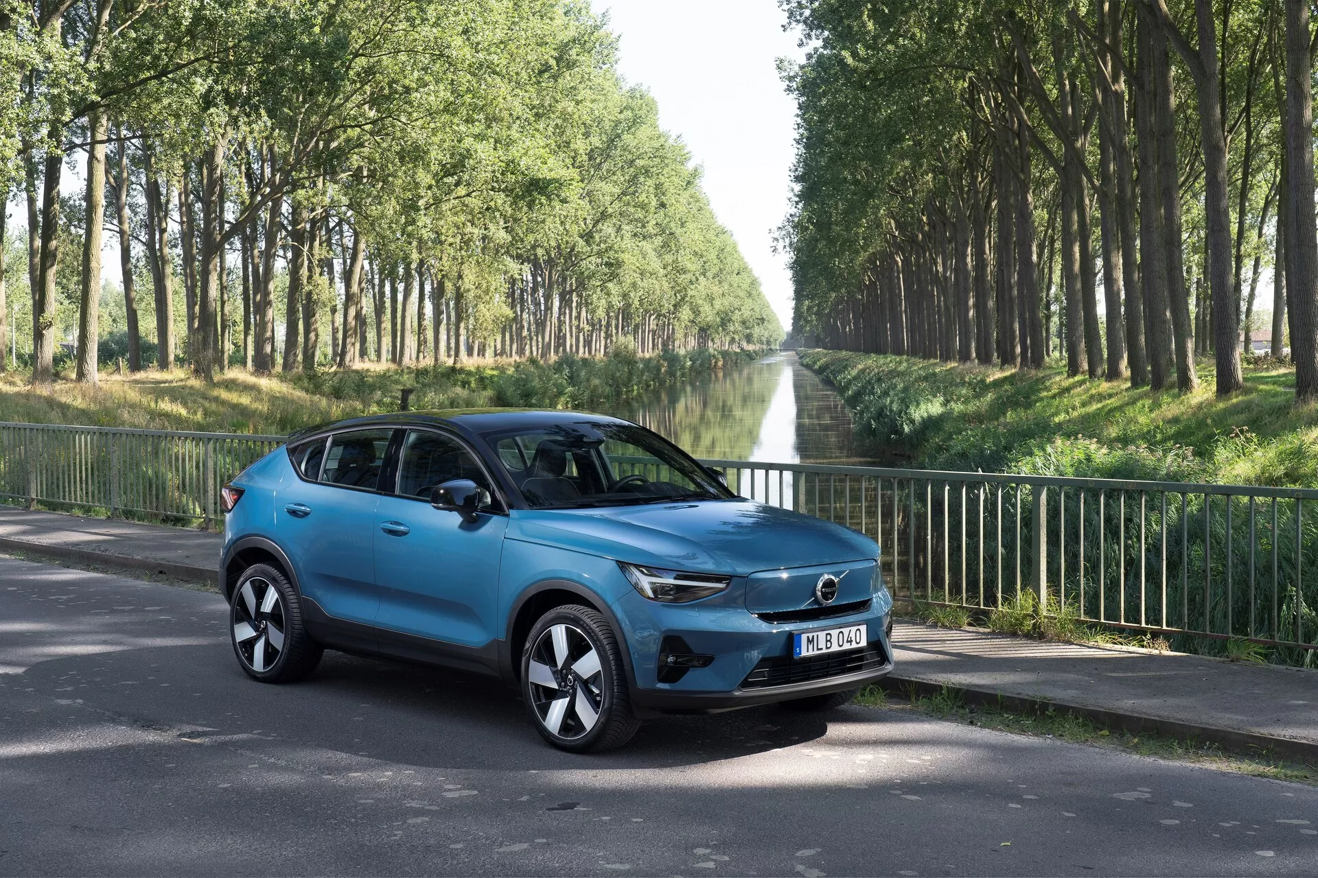 Volvo C40 2021 (prvá generácia) kompaktné SUV v trojštvrťovom pohľade z prednej ľavej strany na asfaltovej ceste. Predstavuje moderné dizajnové prvky ako výraznú masku s centrálnym logom, elegantné LED svetlomety, štýlové zliatinové kolesá, bočné zrkadlá s integrovanými ukazovateľmi smeru, aerodynamické tvarovanie karosérie v jasne modrej farbe, tónované okná a štyri dvere. Umiestnené v vonkajšom prostredí s cestou lemovanou stromami, zamerané na estetické a technologické vlastnosti vozidla.
