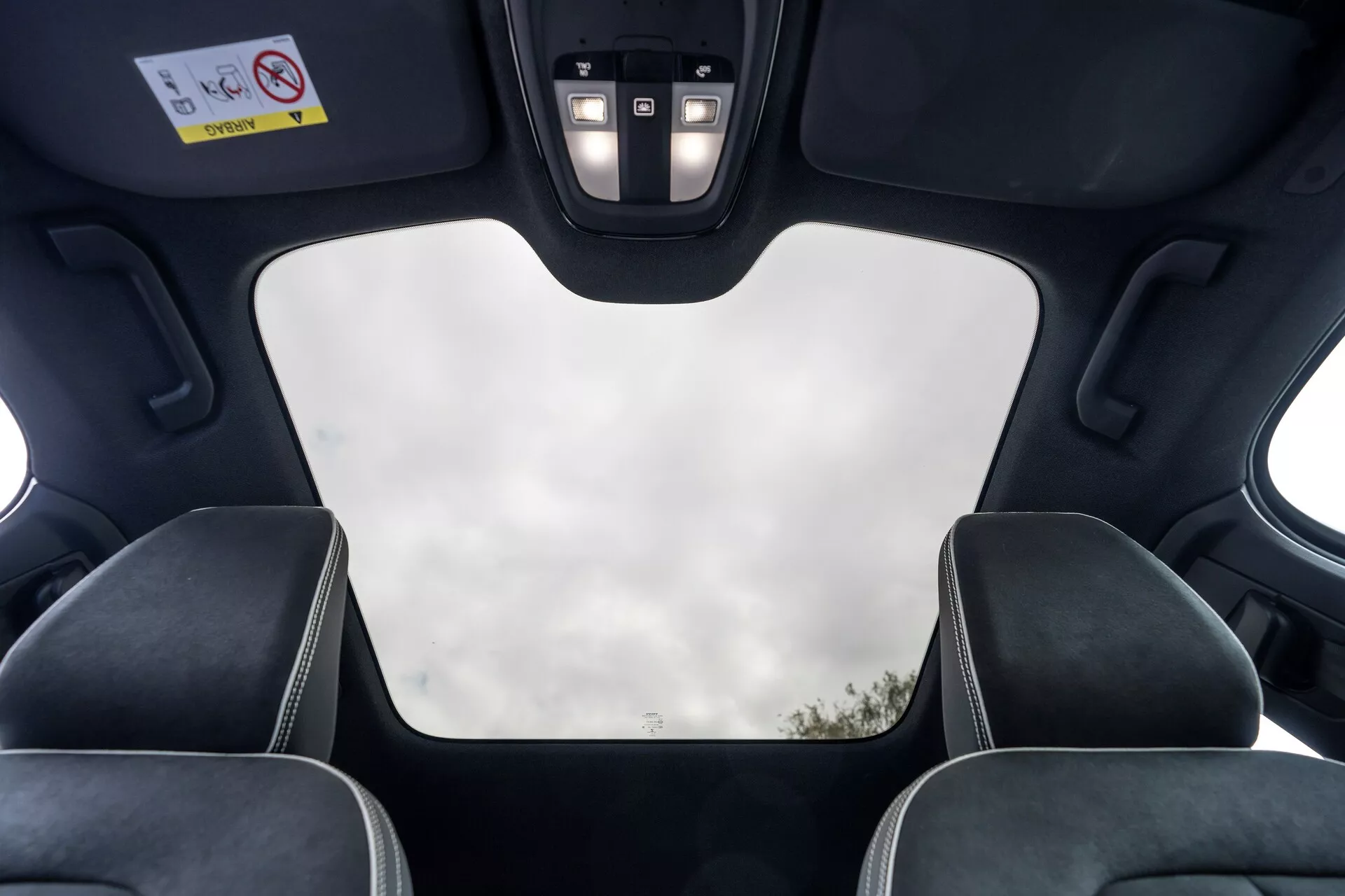 Interiérový pohľad na Volvo C40 2021 (prvá generácia) z perspektívy zadného pasažiera, ktorý ukazuje strešné prvky ako zatvorenú panoramatickú strechu s výhľadom na nebo, čierny obklad, výrezy pre strešný mechanizmus, LED osvetlenie a ovládací panel s tlačidlami pre rôzne funkcie. Obraz tiež zdôrazňuje horné časti hlavových opierok predných sedadiel s kontrastným prešívaním, čo zdôrazňuje moderný luxusný dizajn vozidla a špičkové funkcie.
