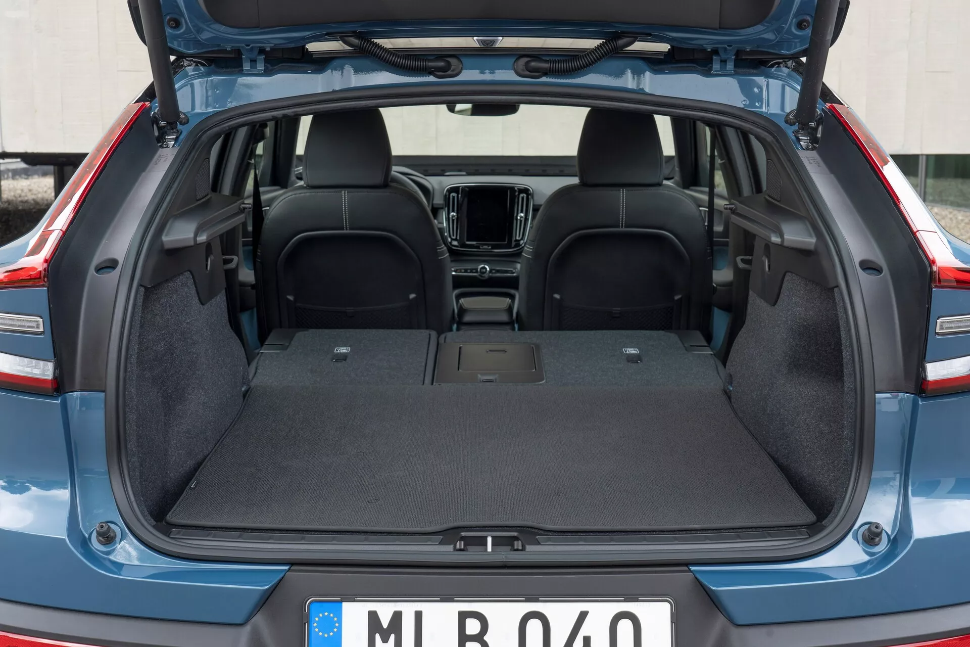 Zadný interiér vozidla Volvo C40 2021 (1. generácia) s otvoreným kufrom, ktorý zobrazuje prázdny batožinový priestor s odolným kobercom, rovným povrchom a bez viditeľných prekážok. Viditeľná je horná časť zadných sedadiel z tmavej látky, s čiastočným pohľadom na prvky prednej kabíny, ako je stredová konzola a infotainmentový systém. Veko kufra je zdvihnuté, čím odhaluje plastové/kompozitné lemovanie okrajov.