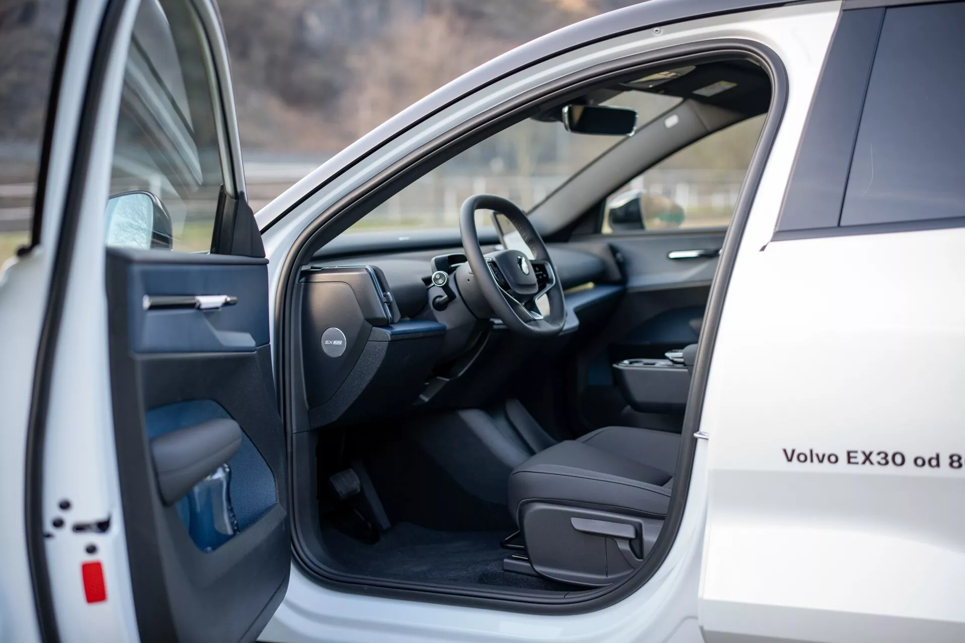 Vnútorný pohľad na bielu Volvo EX30 2024 zobrazujúci sedadlo vodiča a oblasť kokpitu s otvorenými prednými ľavými dvermi s elektrickými ovládačmi, a pohľad na volant a časť prístrojovej dosky