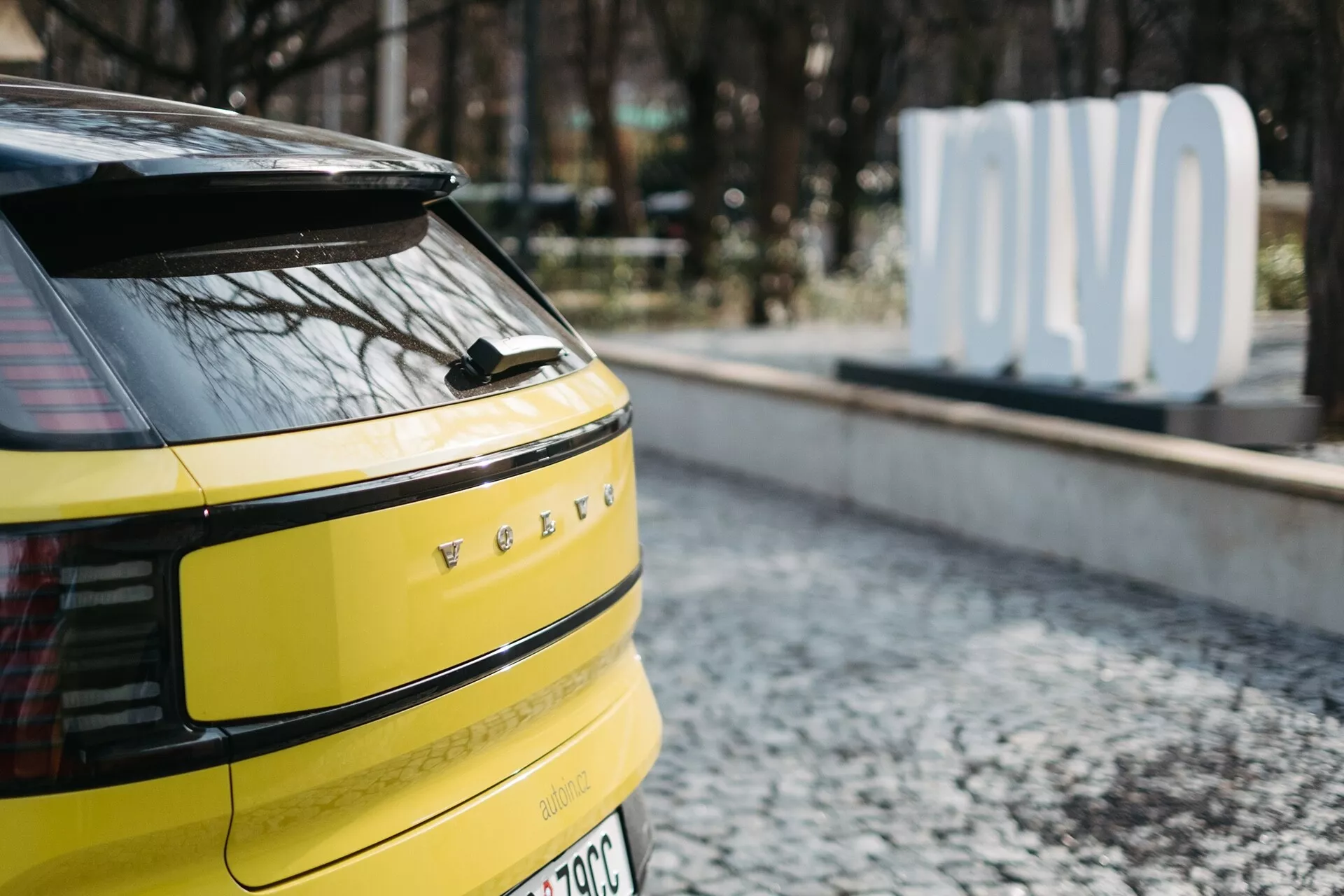 Detailný pohľad na zadnú časť jasne žltého Volvo EX30 2024 so vodorovnými LED zadnými svetlami, potenciálnou zadnou kamerou alebo senzormi, jedným stieračom zadného okna, označením EV alebo hybridu a anténou v tvare žraločej plutvy, zaparkované na povrchu s dlaždicami so zamlženým nápisom 'OLVO' na pozadí