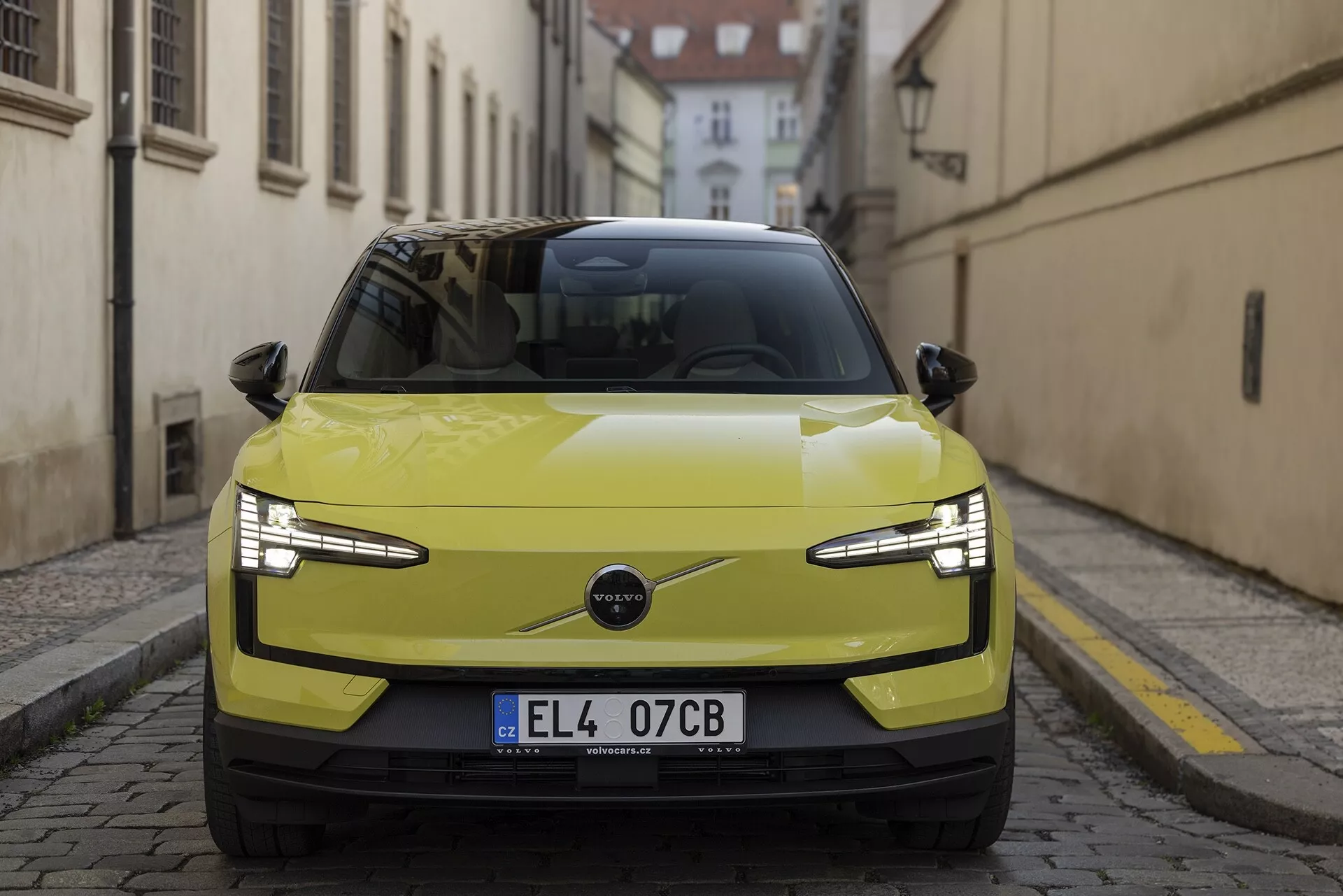 Predný pohľad na Volvo EX30 2024 v limetkovo žltej farbe, ktorý zdôrazňuje jeho štýlové LED svetlomety, minimalistickú mriežku naznačujúcu elektrický pohon a pokročilé technologické vlastnosti ako parkovacie senzory a možné ADAS, proti historickému európskemu ulici