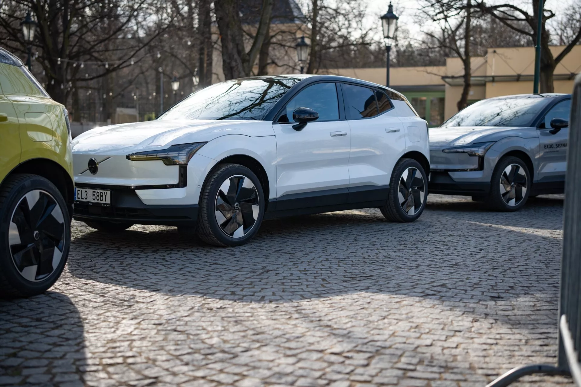Biely moderný crossover SUV model 2024 v trojštvrťovom pohľade zaparkovaný na dlaždenej ulici, s lesklým lakovaním, LED dennými svetlami, zapustenými kľučkami dverí a dynamicky navrhnutými zliatinovými kolesami naznačujúcimi luxus a výkon, pravdepodobne elektrické vozidlo.