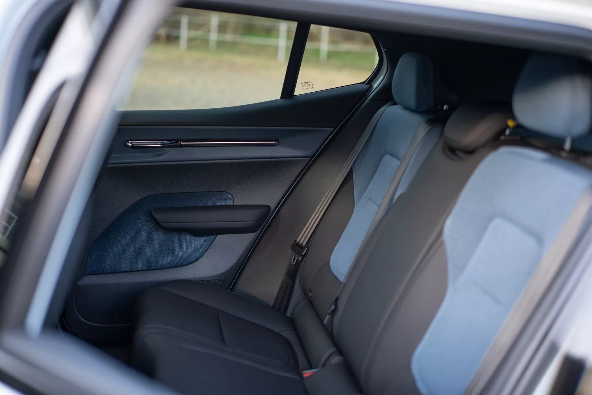 Interiér Volvo EX30 2024 zobrazujúci zadnú pasažiersku časť cez otvorené dvere spolujazdca vpredu, s tmavým čalúnením, viditeľnými opierkami hlavy, detailným panelom dverí s ovládacími prvkami, dizajnom B-stĺpika, bezpečnostnými pásami na zadných sedadlách, čiastočne viditeľným bočným oknom zlepšujúcim atmosféru interiéru a zameraním na úpravu sedadiel pre pohodlie a bezpečnosť pasažierov.