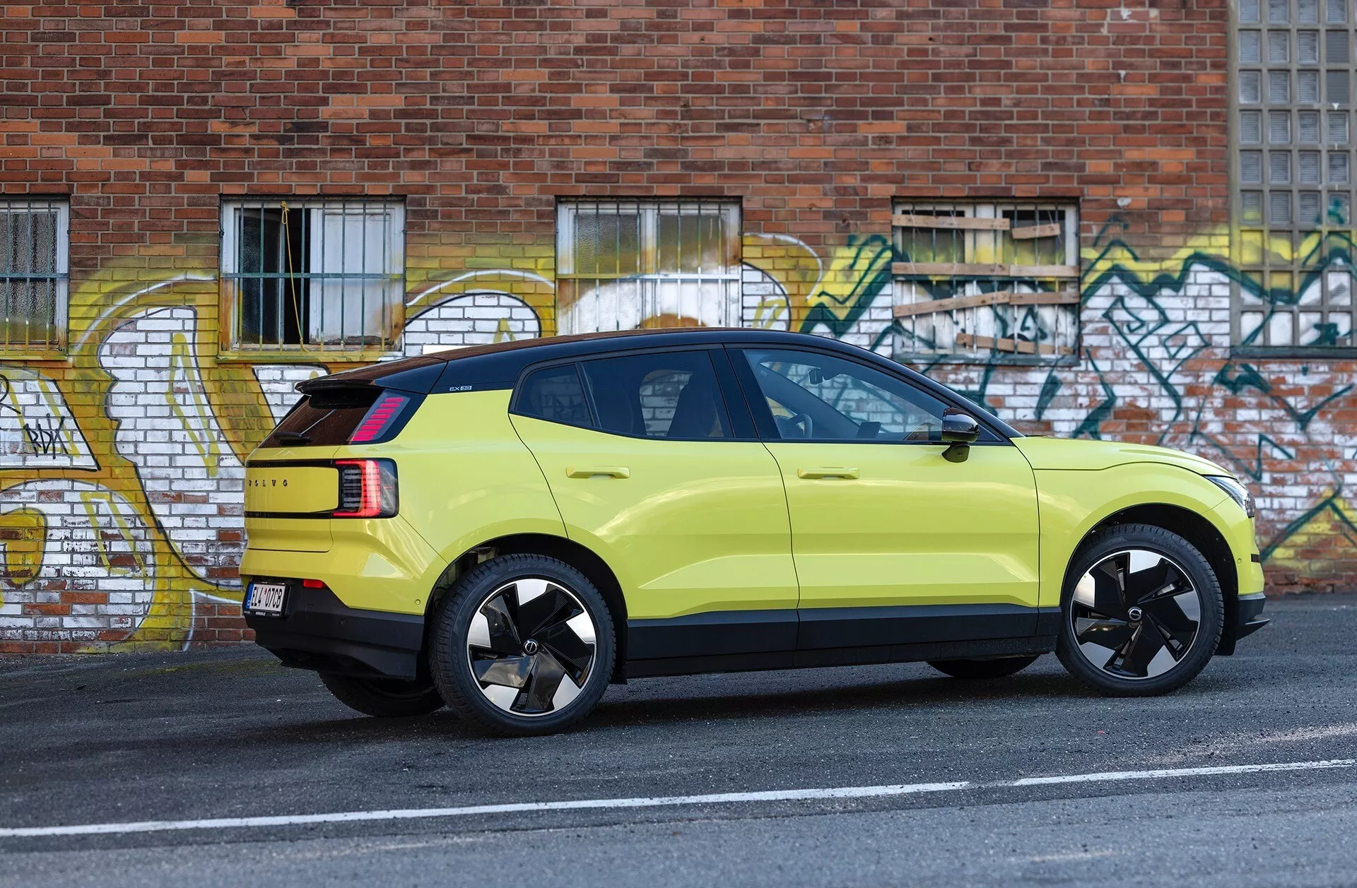 Bočný pohľad na limetkovo zelené SUV Volvo EX30 ročník 2024 zaparkované vedľa obrubníka mestské ulice s grafitovým umením na pozadí, predvádzajúce športové čierne zliatinové disky, LED zadné svetlá a vysoko umiestnenú brzdovú lampu.