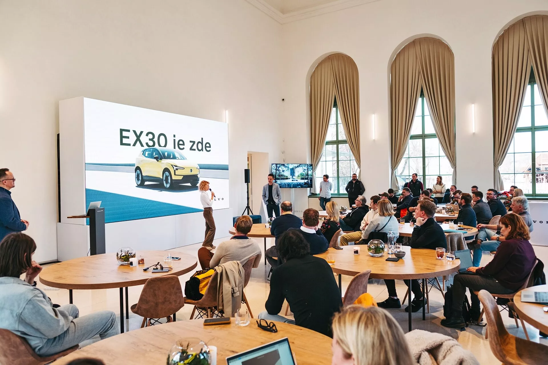 Prezentácia elektrického vozidla Volvo EX30 2024 v priestrannej miestnosti s vysokými stropmi a vysokými oblúkovými oknami, kde je zobrazené žlté EX30 z trojštvrťového predného pohľadu na veľkej obrazovke