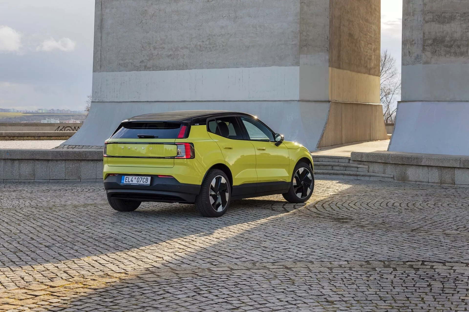 Limetkově zelené SUV Volvo EX30 2024 v zobrazení z troch štvrtín zozadu, predstavujúce bočný profil, zadné svetlá, veko batožinového priestoru a čierne zliatinové kolesá pri kameňových múroch na dlažobnom povrchu