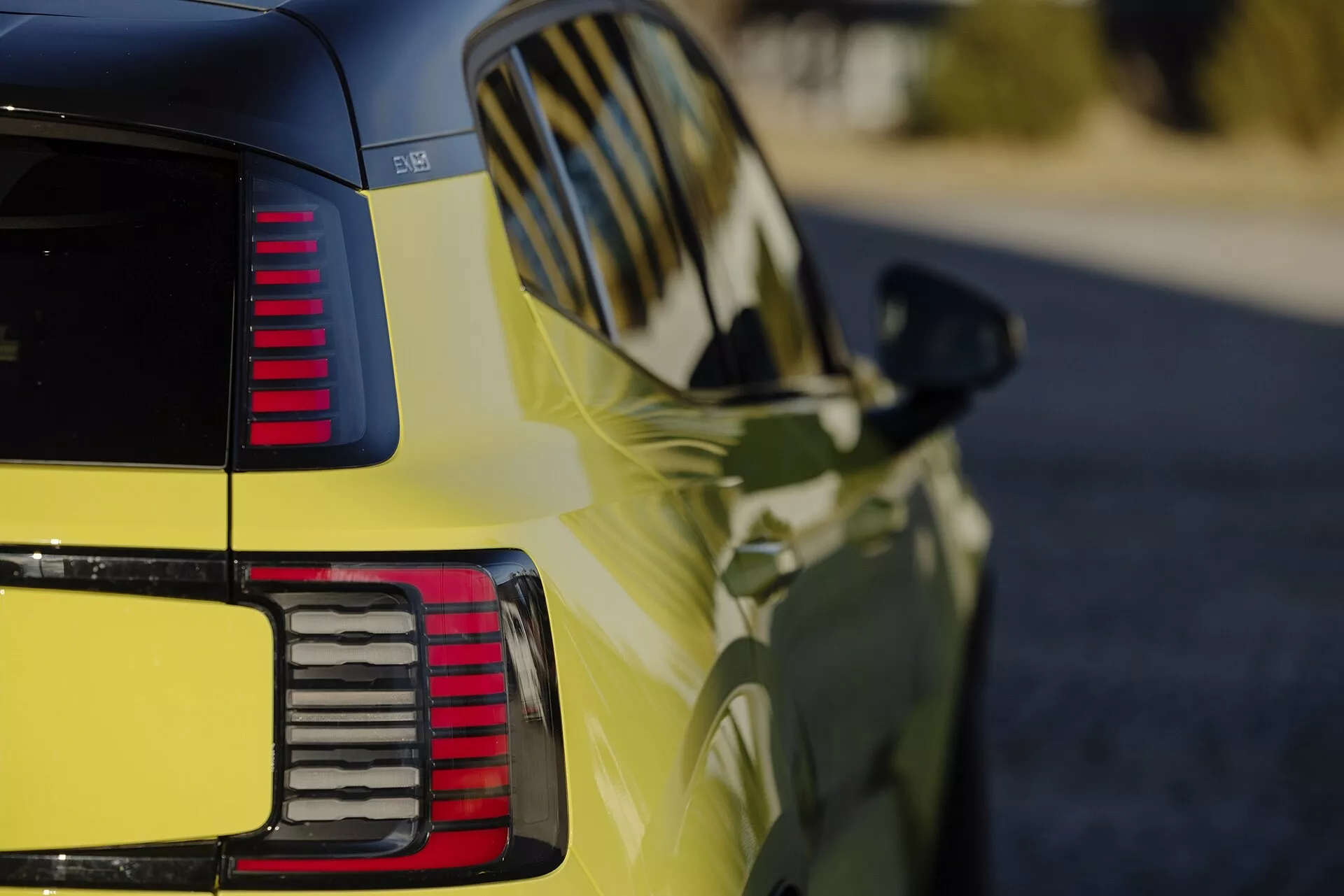 Detailný pohľad na zadnú štvrtinu Volvo EX30 2024 vo vibrantnej žltej, ktorá zobrazuje lesklý povrch, segmentované LED zadné svetlá, aerodynamické dizajnové línie a integrované smerové signály na bočnom zrkadle