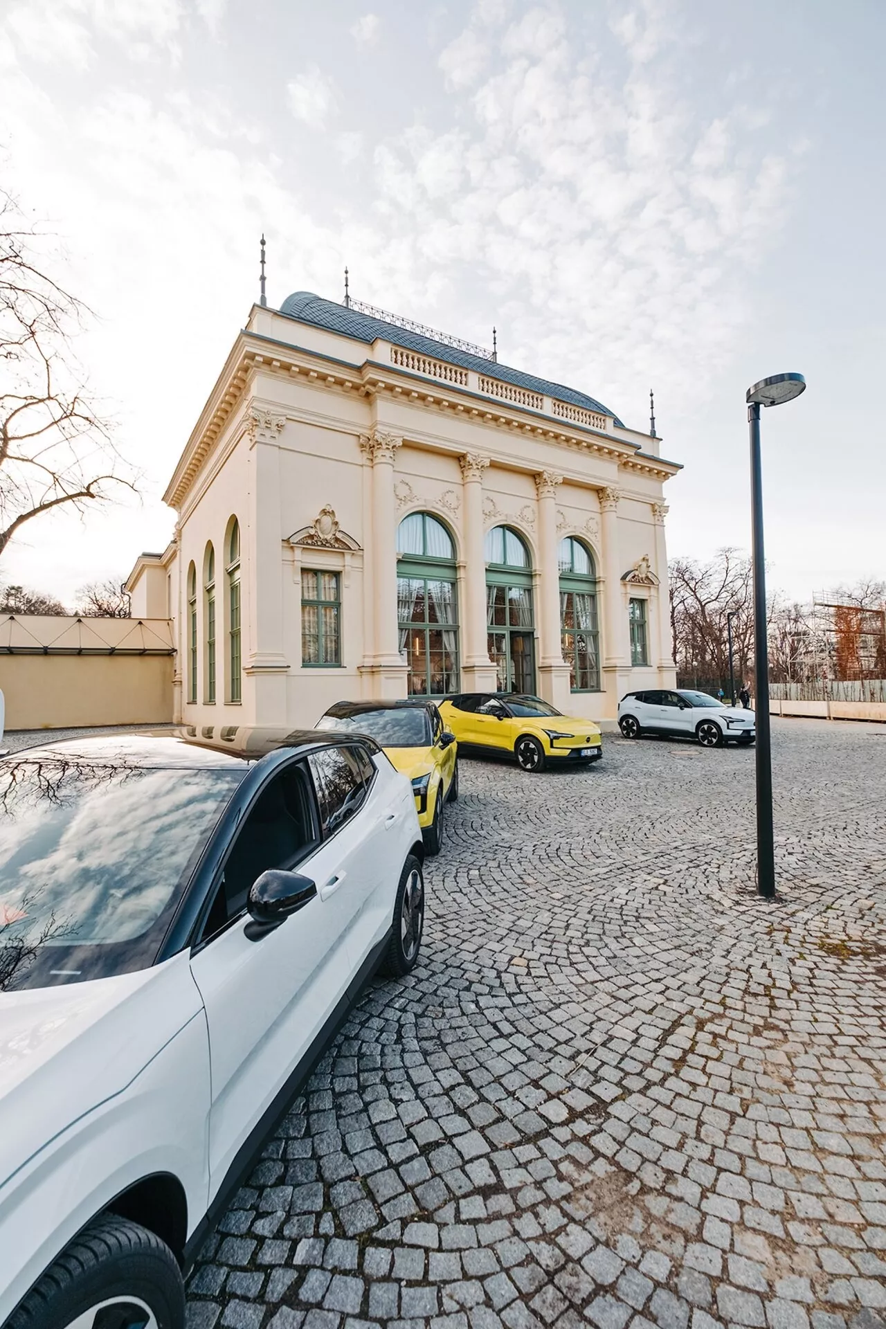Mestská scéna s tromi autami a elegantnou budovou na pozadí, zhotovená z uhla širokouhlého objektívu, zvýrazňujúca biely súčasný sedan, výrazné žlté športové auto a biely sedan alebo hatchback v svetle neskorého popoludnia