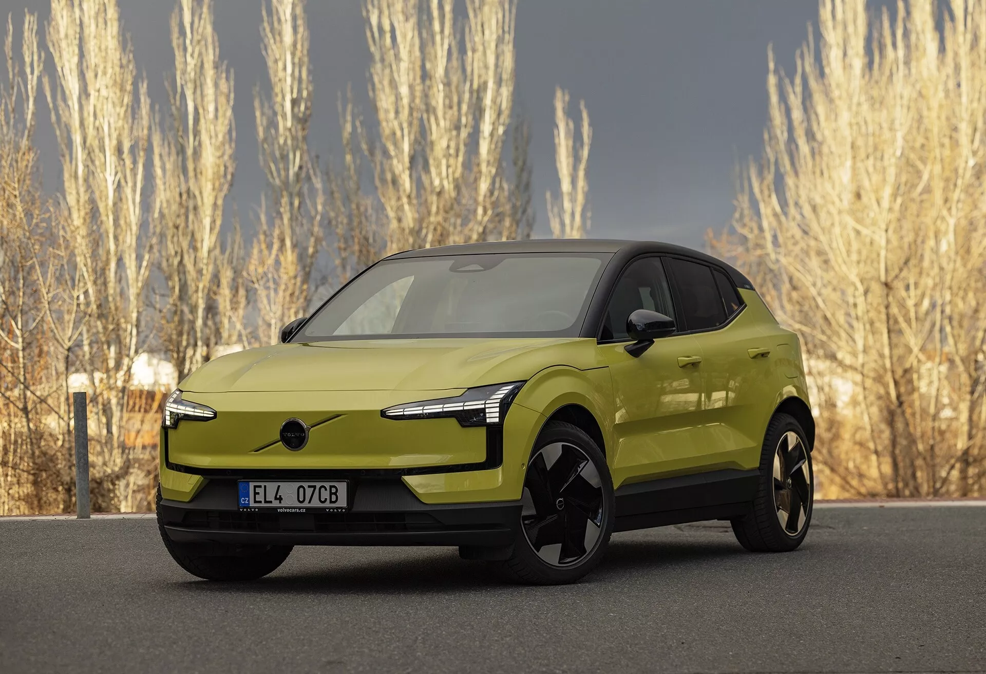 Moderné limetkovo zelené elektrické auto Volvo EX30 2024 vo štvrtinovom prednom pohľade zdôrazňujúce jeho elegantné LED svetlomety, absenciu tradičnej mriežky pre lepšiu aerodynamiku, veľké čierne zliatinové kolesá, vonkajšie zrkadlá vo farbe karosérie s potenciálne integrovanou technológiou, hladký a aerodynamický bočný profil s vzhľadom podobným kupé a živý metalický povrch, zaparkované v prírodnom prostredí