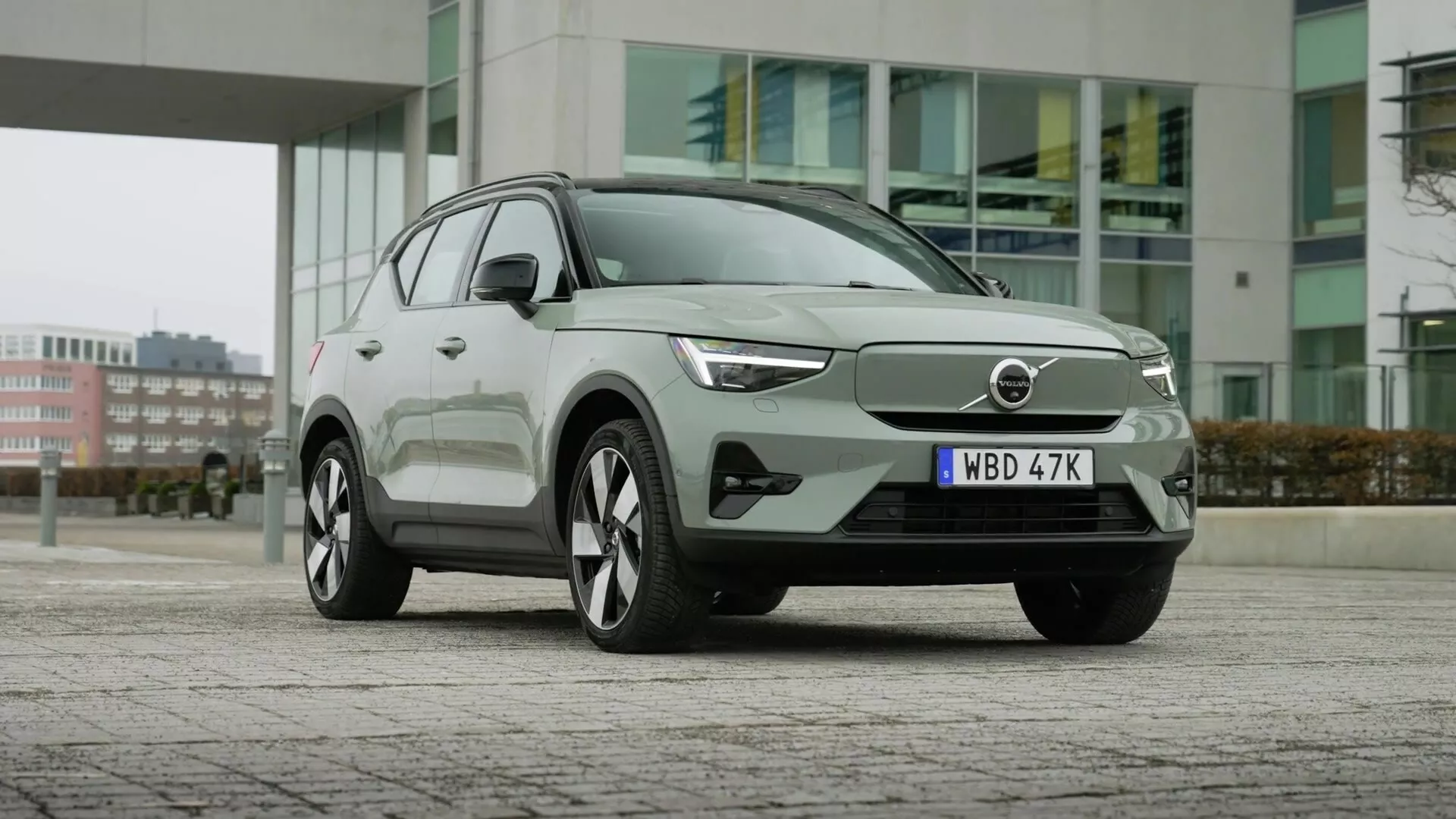 Elektrické SUV Volvo EX40 1. generácie z roku 2024 zaparkované na dláždenom povrchu s mestskými budovami na pozadí, predvádzajúce svoj aerodynamický predný diel s uzavretou mriežkou, elegantné LED svetlomety, veľké viacramenné zliatinové kolesá, moderný dizajn karosérie s úrovňovými kľučkami dverí, integrované smerové signály v bočných zrkadlách, okná s chrómovým rámom a pokročilú technológiu asistencie vodiča.