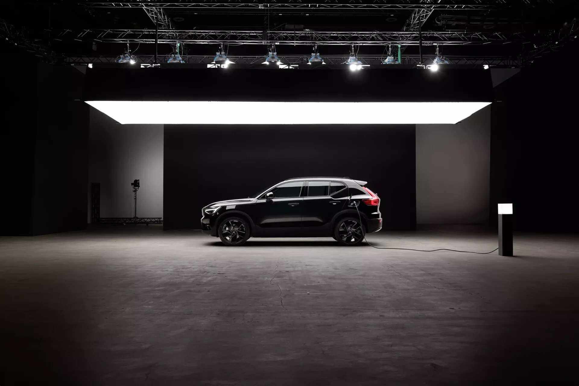 Bočný profil Volvo EX40 1. generácie pre rok 2024 v štúdiovom prostredí, predstavuje jeho elegantný čierny exteriér, moderný dizajn SUV so športovým crossover štýlom a možnosti elektrického/hybridného pohonu. Funkcie zahŕňajú veľké kolesá s dizajnom z ľahkej zliatiny s viacerými ramenami, dramatické osvetlenie bez odrazu a nabíjací kábel naznačujúci technológiu udržateľnej mobility.