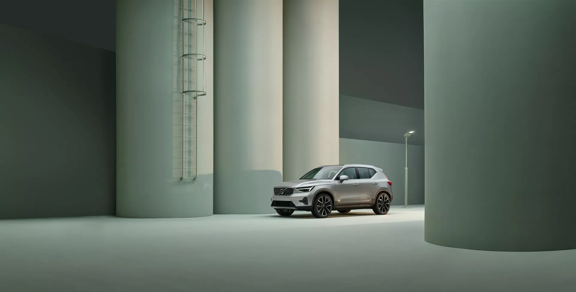 Obrázok zobrazuje Volvo XC40 2022 (prvá generácia facelift) v priemyselnom alebo architektonickom prostredí s veľkými valcovými štruktúrami. Zachytený z trojštvrťového predného uhla, zdôrazňuje moderný dizajn vozidla, vrátane ostrých LED svetiel a výraznej mriežky. Priestranné prostredie a mäkké osvetlenie zdôrazňujú prémiové vlastnosti, ako je pokročilá osvetľovacia technológia, vysokokvalitný lak a sofistikované kolesá, ktoré naznačujú výkonnostnú alebo luxusnú verziu.