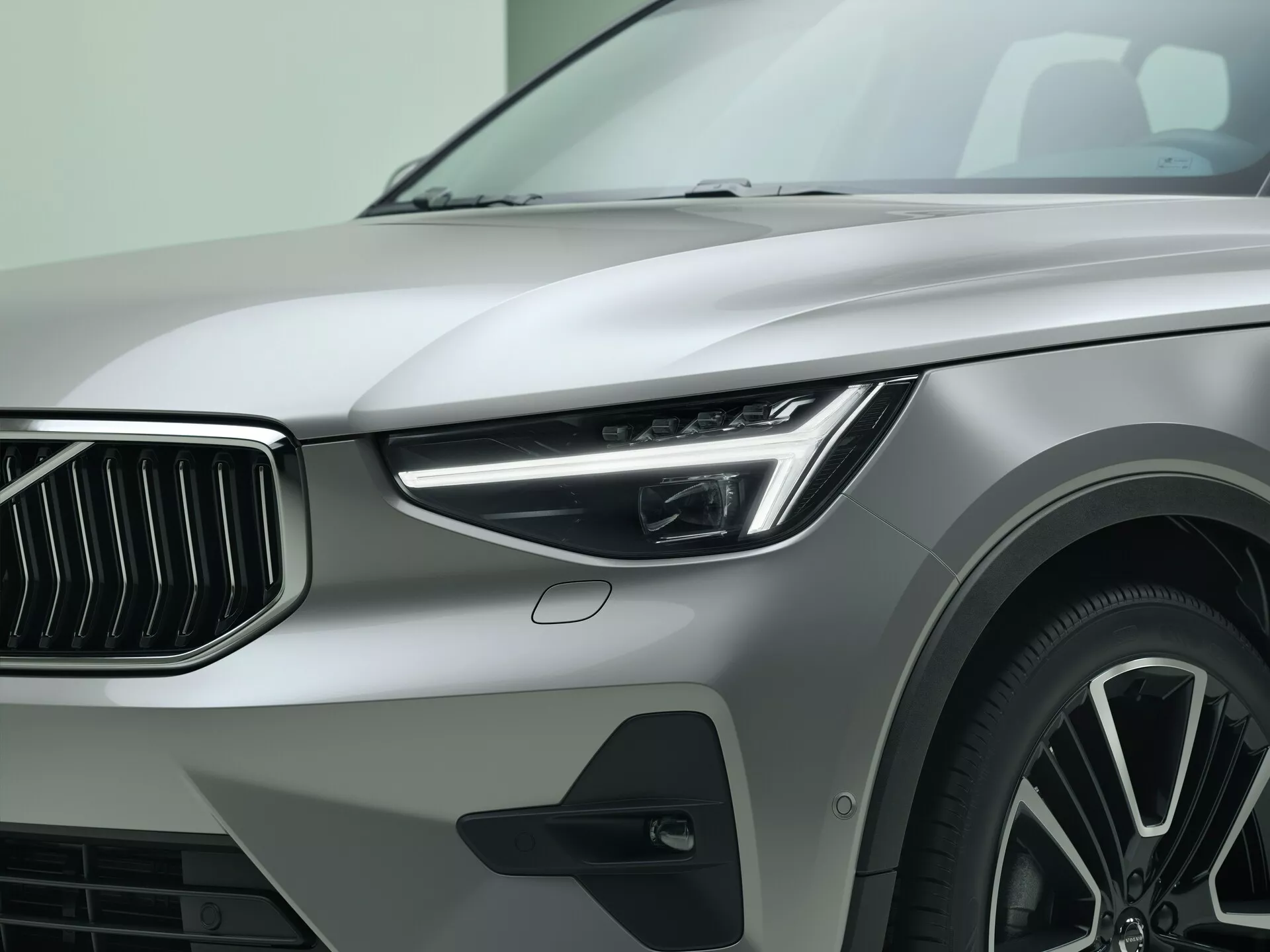 Detailný pohľad na ľavú prednú štvrť vozidla Volvo XC40 2022 prvej generácie facelift, zobrazujúci elegantný LED svetlomet s denným svietením, výraznú prednú mriežku so zvislými lamelami, vysokovýkonnú nízkoprofilovú pneumatiku na viaclúčovom zliatinovom kolese, metalický lak a ďalší dizajnový prvok blatníka, zdôrazňujúci luxus a moderný dizajn automobilov.