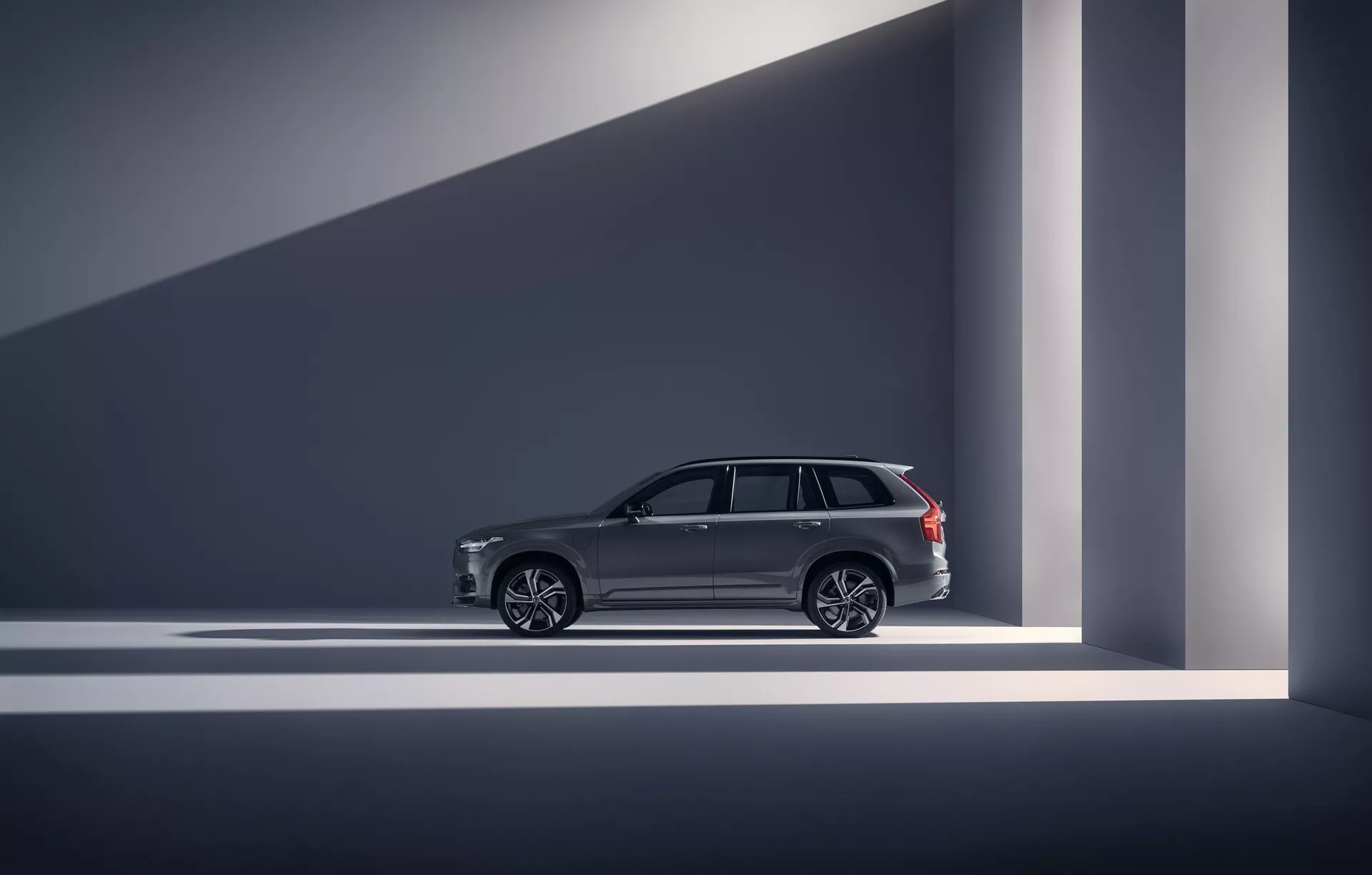 Bočný profil vozu 2015 Volvo XC90 (2. generácia) v kategórii luxusných SUV. Fotografia je odfotená v interiérovom štúdiu alebo showroomu, zobrazujúca dizajnové línie vozidla, elegantné kľučky dverí, veľké viacramenné zliatinové kolesá a chrómové obloženie okien. Strešné nosiče naznačujú kombináciu úžitkovosti a luxusu. Vozidlo je dobre osvetlené dramatickým osvetlením, ktoré vytvára geometrické tiene a zdôrazňuje jeho moderný a vysoko kvalitný vzhľad. Pokročilé funkcie ako napríklad bezkľúčový vstup sú naznačené dizajnovými prvkami.