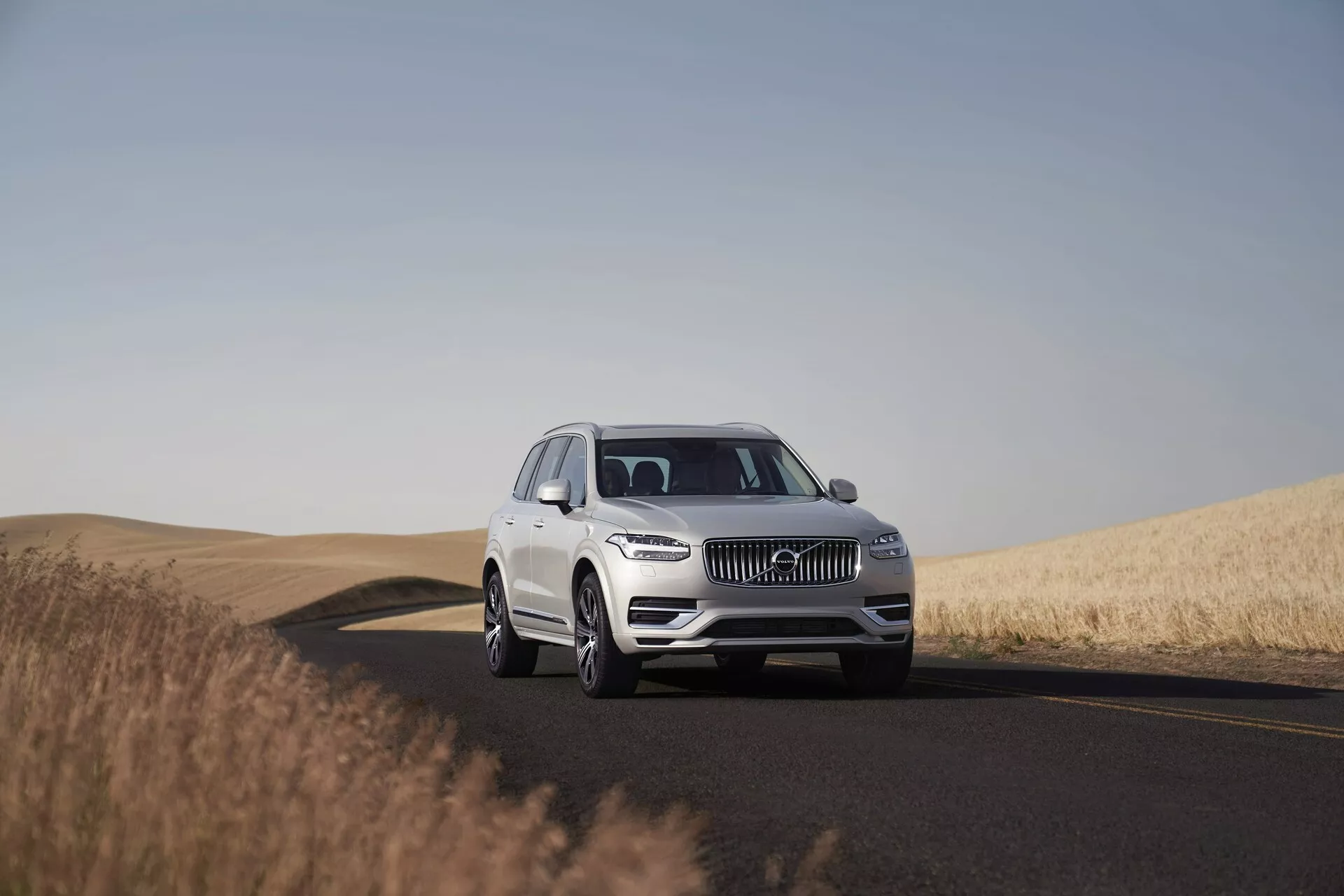 Biele SUV Volvo XC90 z roku 2015 na dvojpruhovej ceste, zachytené počas denného svetla s jasnou oblohou. Obrázok ukazuje vozidlo z nízkeho až stredného uhla, zvýrazňujúc jeho impozantnú prítomnosť a rozmery. Umiestnené v strede rámu s trojštvrťovým pohľadom, fotografia zachytáva detaily dizajnu ako mriežku, LED svetlomety, emblém a predný nárazník, indikujúce pokročilé asistenčné systémy pre vodiča. Kolesá, zliatiny a prípadne veľkosť pneumatík sú jasne viditeľné. Pokojné prostredie obsahuje cestu zatáčajúcu doprava a zlaté pšeničné polia, zdôrazňujúc vhodnosť vozidla pre dlhé cesty v rozmanitých krajinách.