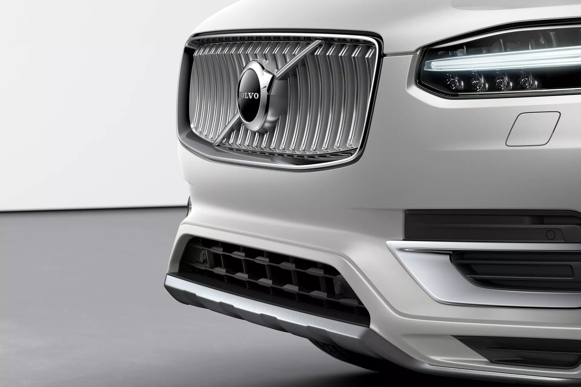 Detailný záber prednej časti bieleho Volvo XC90 2015 druhej generácie so zvýraznenou mriežkou s vertikálnymi lamelami a charakteristickým emblémom Volvo v strede. Chrómová úprava mriežky kontrastuje s matnou textúrou okolo nej. Viditeľné sú LED denné svetlá Thorovo kladivo v hlavnom svetlomete. Spodný nárazník obsahuje veľký prívod vzduchu s ozdobným strieborným lemom, čo naznačuje aerodynamickú účinnosť a chladenie motora. Záber z mierne nižšieho uhla zdôrazňuje eleganciu, kvalitu spracovania a prémiový predný dizajn značky Volvo.