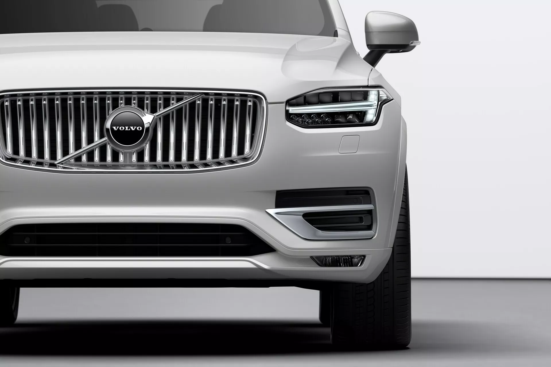 Záber zblízka na prednú štvrtinu strieborného vozidla Volvo XC90 z roku 2015 (2. generácia) zhotovený z nízkeho uhla. Vyznačuje sa výraznou chrómovou mriežkou s charakteristickým znakom Volvo, elegantnými svetlometmi s dennými LED svetlami v tvare T, označovanými výrobcom ako 'Thorovo kladivo', a dobre integrovanými parkovacími senzormi na prednom nárazníku. Spodná časť nárazníka obsahuje nasávacie otvory a kryty hmlových svetiel orámované strieborným lemovaním. Viditeľné vonkajšie zrkadlá naznačujú pokročilé technologické prvky ako smerovky alebo systémy varovania pred mŕtvym uhlom.
