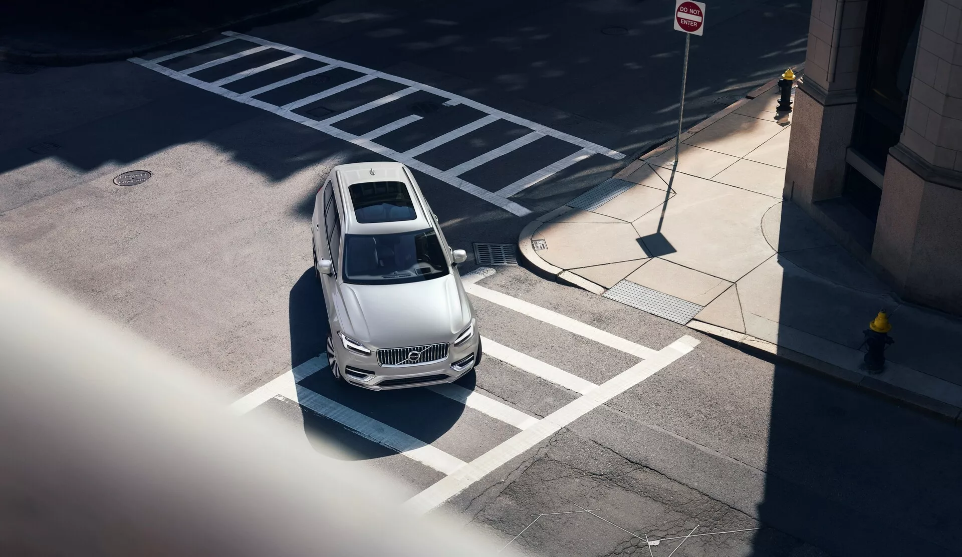 Pohľad zhora na Volvo XC90 z roku 2015 (2. generácia) z vysokého uhla, diagonálne od predného ľavého rohu; zobrazuje svetlú kapotu auta, strechu a časť vodičovej strany. Auto je správne zaparkované na vyznačenom mieste blízko prechodu pre chodcov a obsahuje dizajnové prvky naznačujúce aerodynamiku, čisté čelné sklo odhaľujúce čiastočne viditeľný interiér, výrazné strešné okno a ostré bočné línie. Mestské prostredie je zdôraznené blízkymi parkovými a dopravnými značkami, s výraznými tieňmi, ktoré naznačujú poludňajšie osvetlenie. Vozidlo tiež naznačuje moderné technológické funkcie bez viditeľných senzorov a kamier.